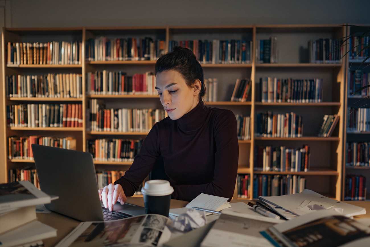 A professional studying alone at night in a library, surrounded by open books and papers