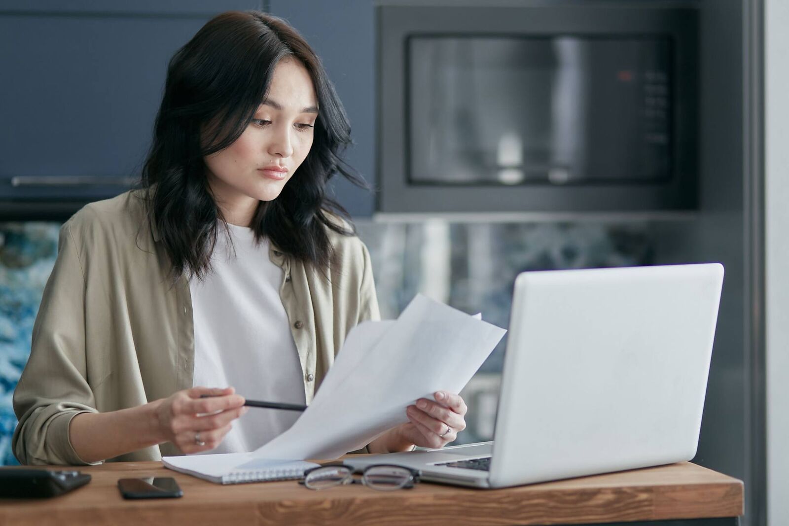 A woman reviews printed pages beside an open laptop at a bright kitchen counter