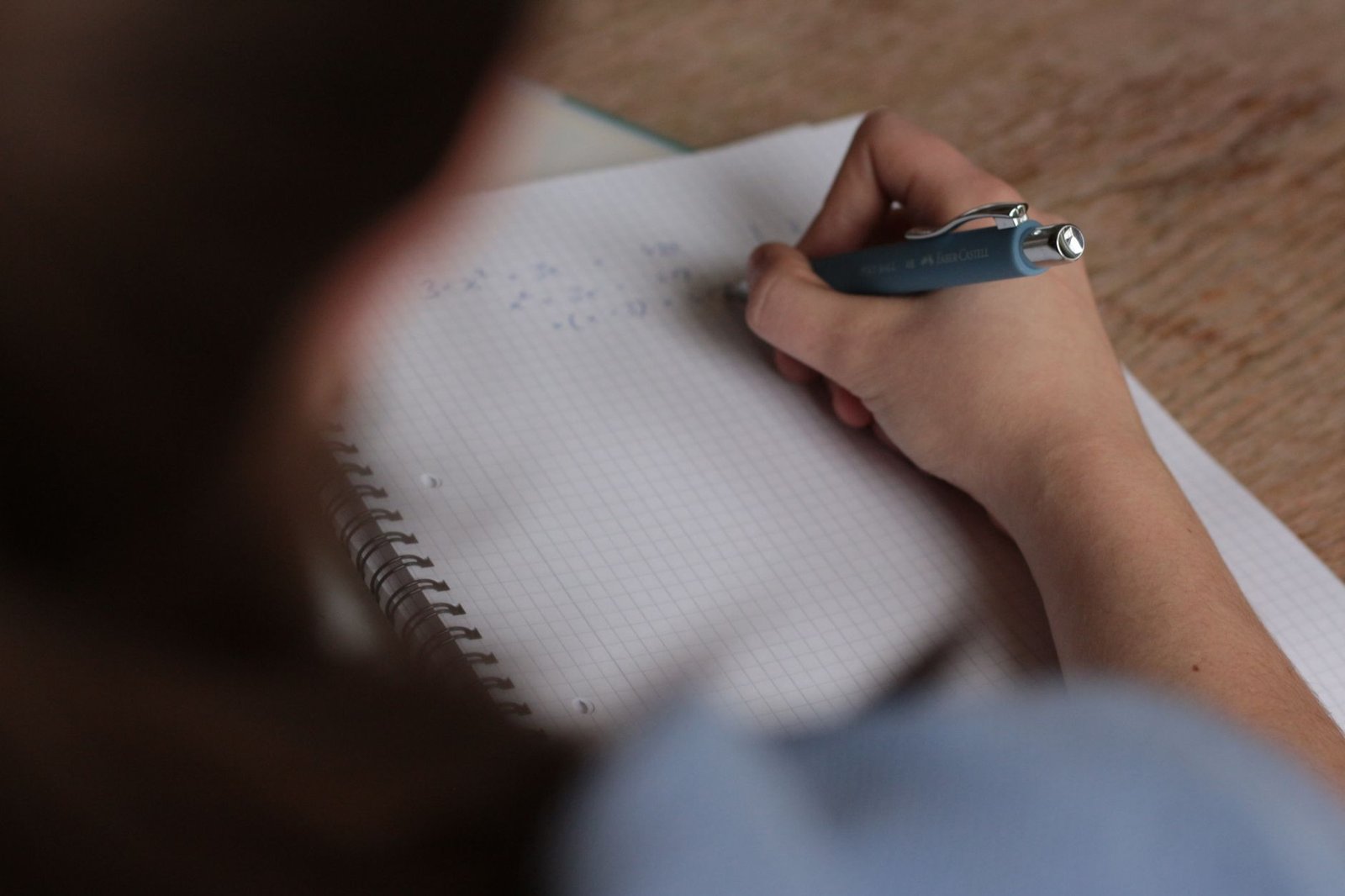 A close-up of a hand annotating handwritten notes in a spiral notebook with a pen, seen over the writer&rsquo;s shoulder