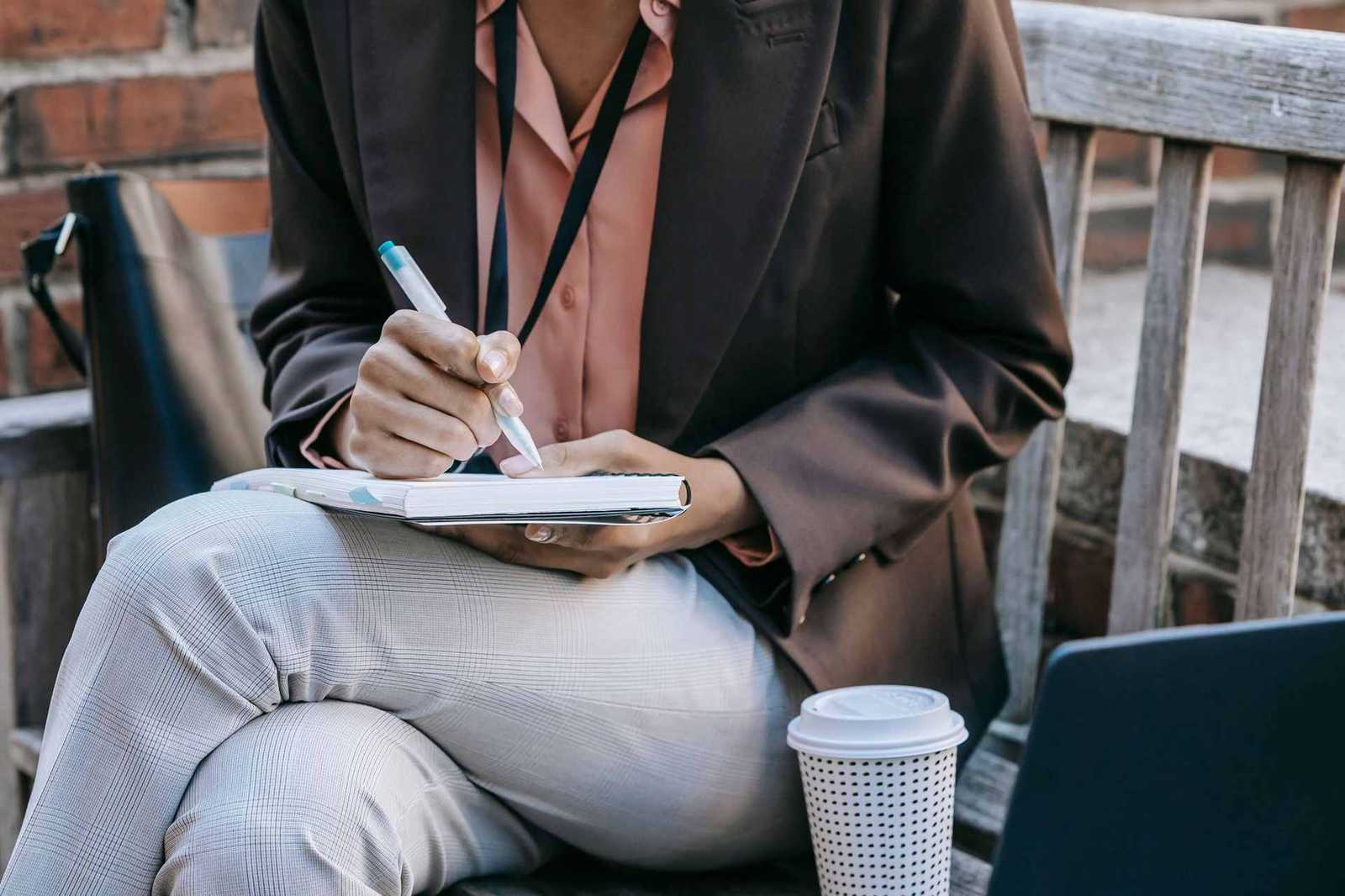 A person writes in a notebook resting on their lap, a lanyard visible at the collar, a coffee cup and laptop nearby