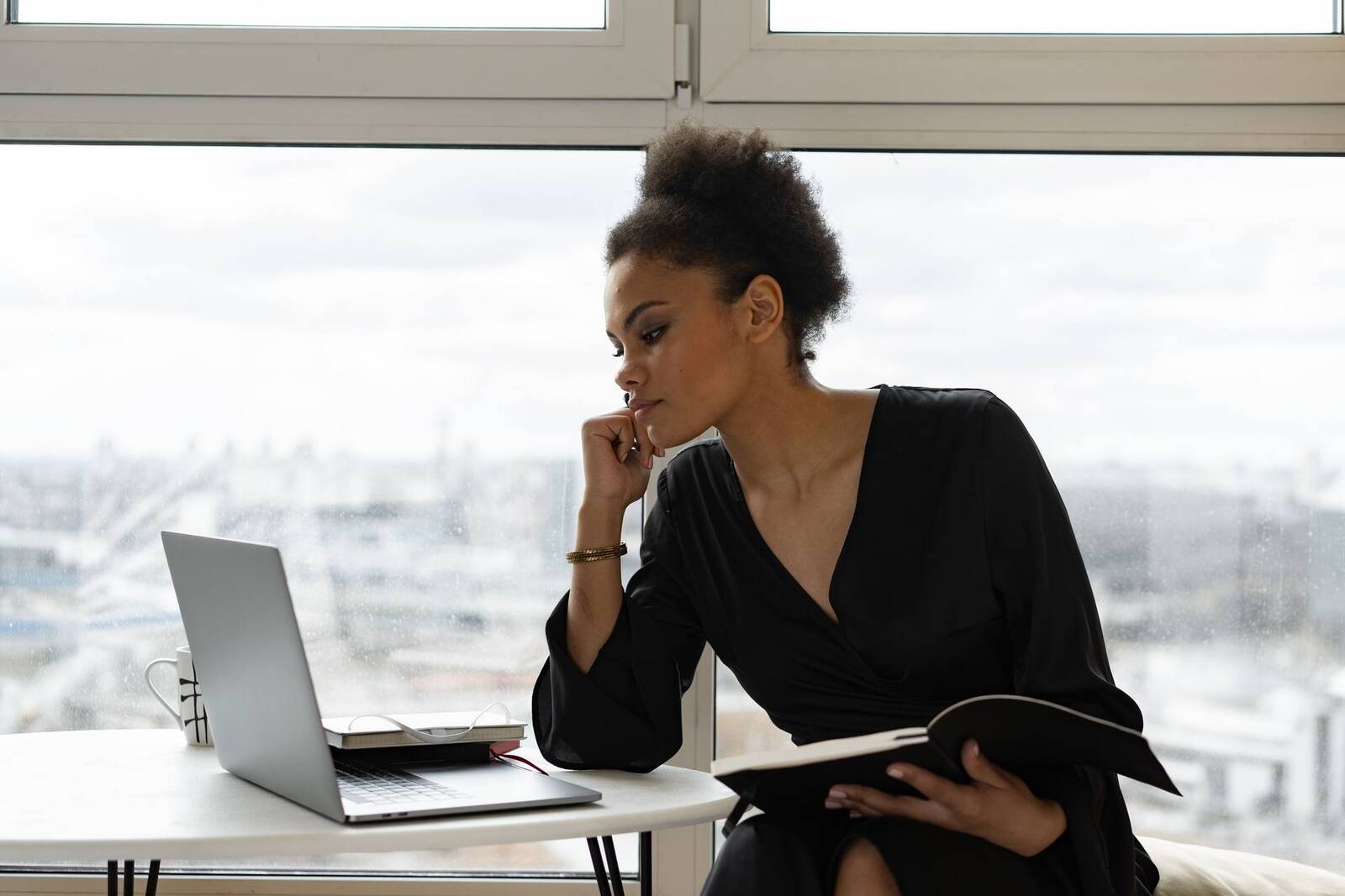 A woman rests her chin on her hand, looking intently at a laptop beside a rain-streaked office window overlooking a city