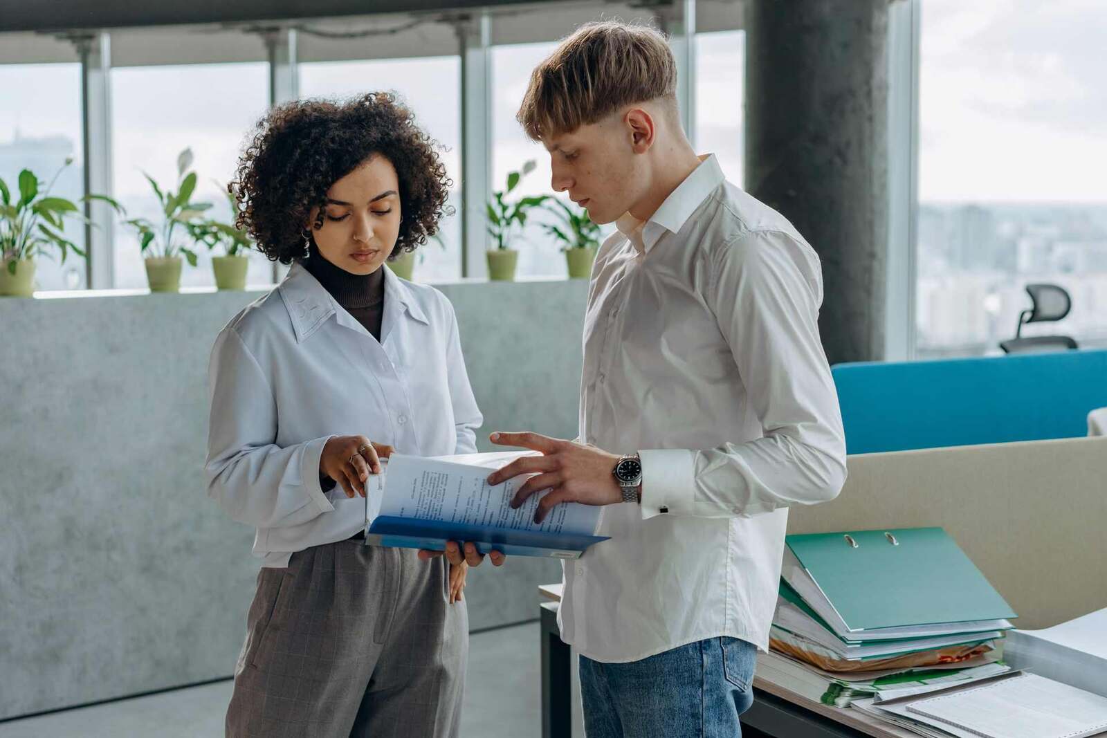 Two colleagues in white shirts stand together reading an open binder of printed pages in a bright office