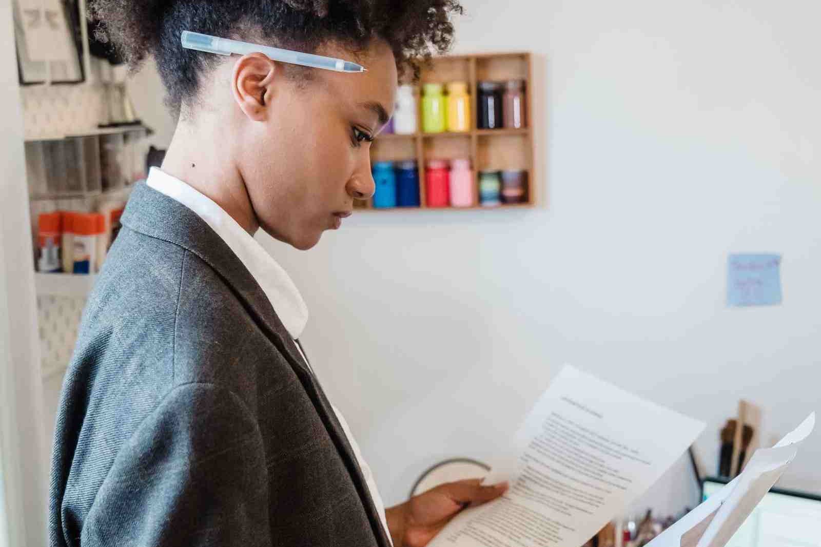 A woman in a blazer reads a printed document closely while standing in a home office, a pen tucked behind her ear