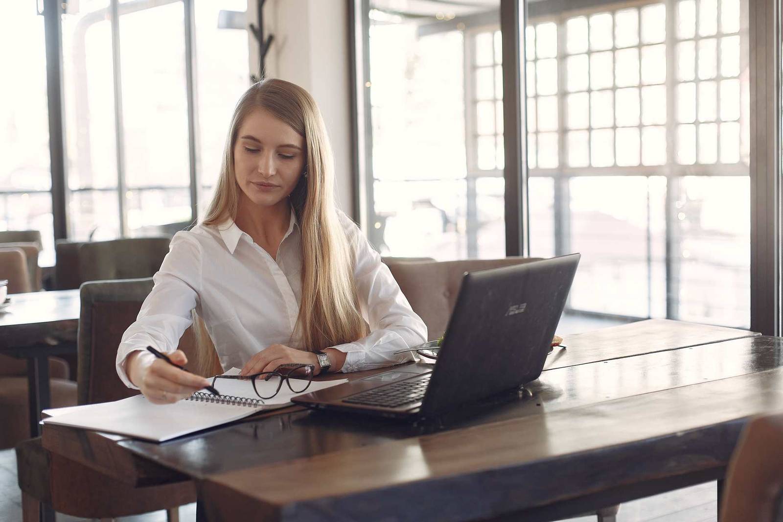 A woman in a white shirt writes in a notebook at a wooden table beside an open laptop