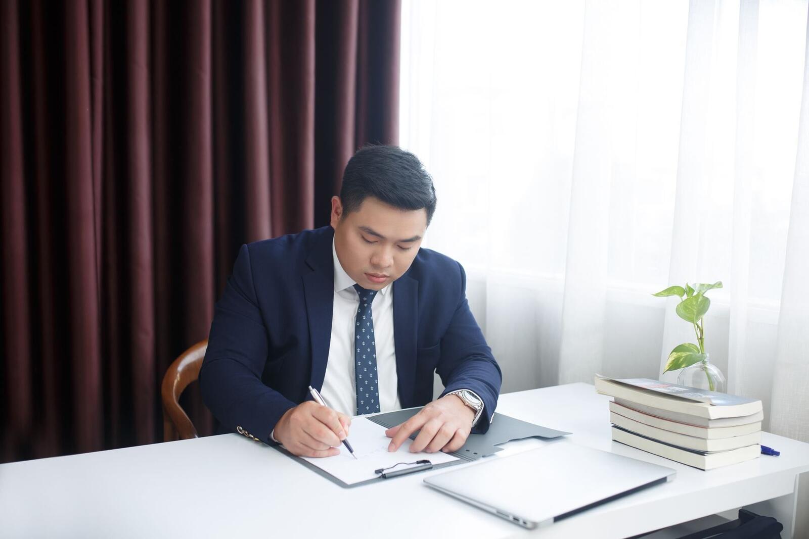 A man in a navy suit writes on a clipboard at a white desk beside a closed laptop, bright curtained window behind him
