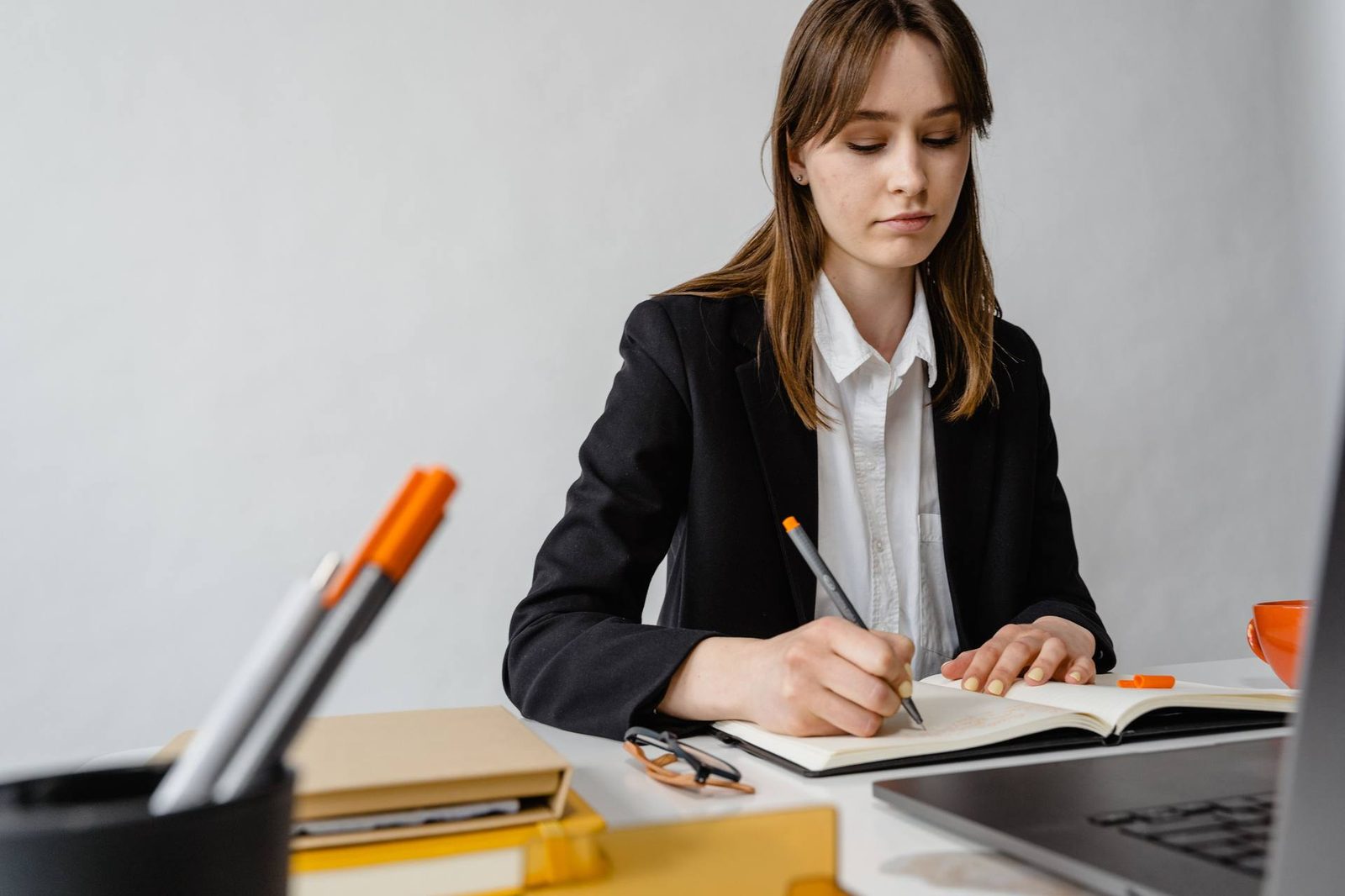 A woman in a blazer writes in a notebook at a desk beside an open laptop, glasses set to one side