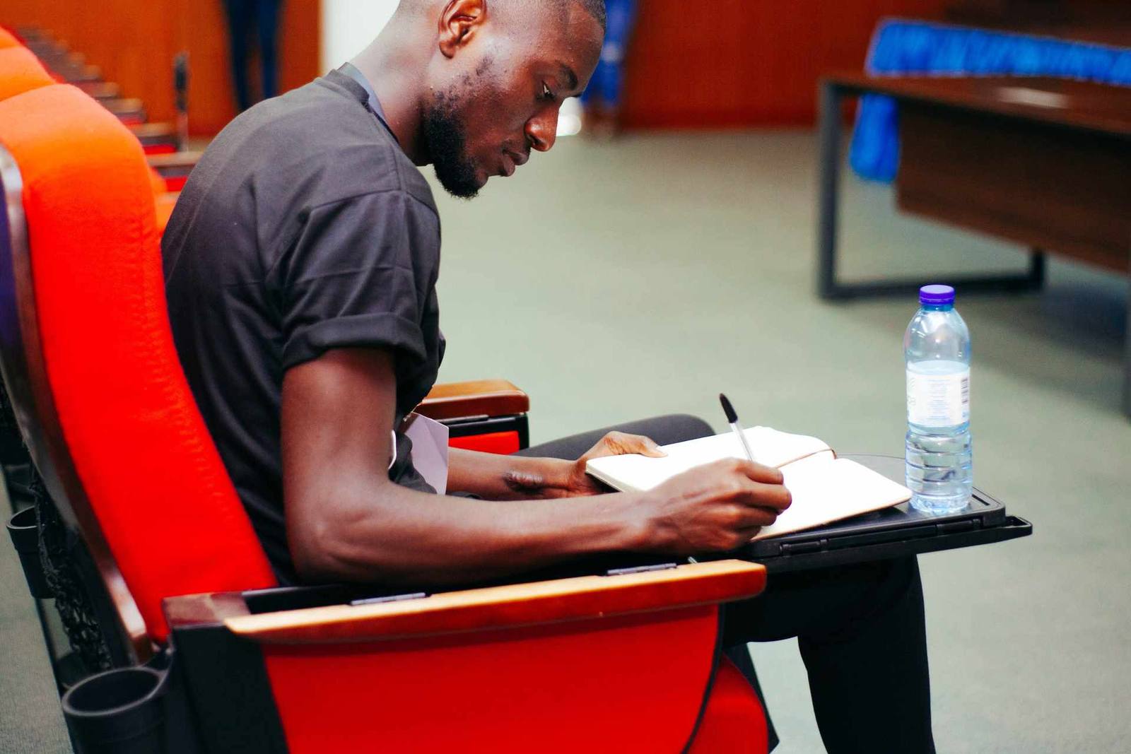 A man writes closely in a notebook in a red-seated lecture hall