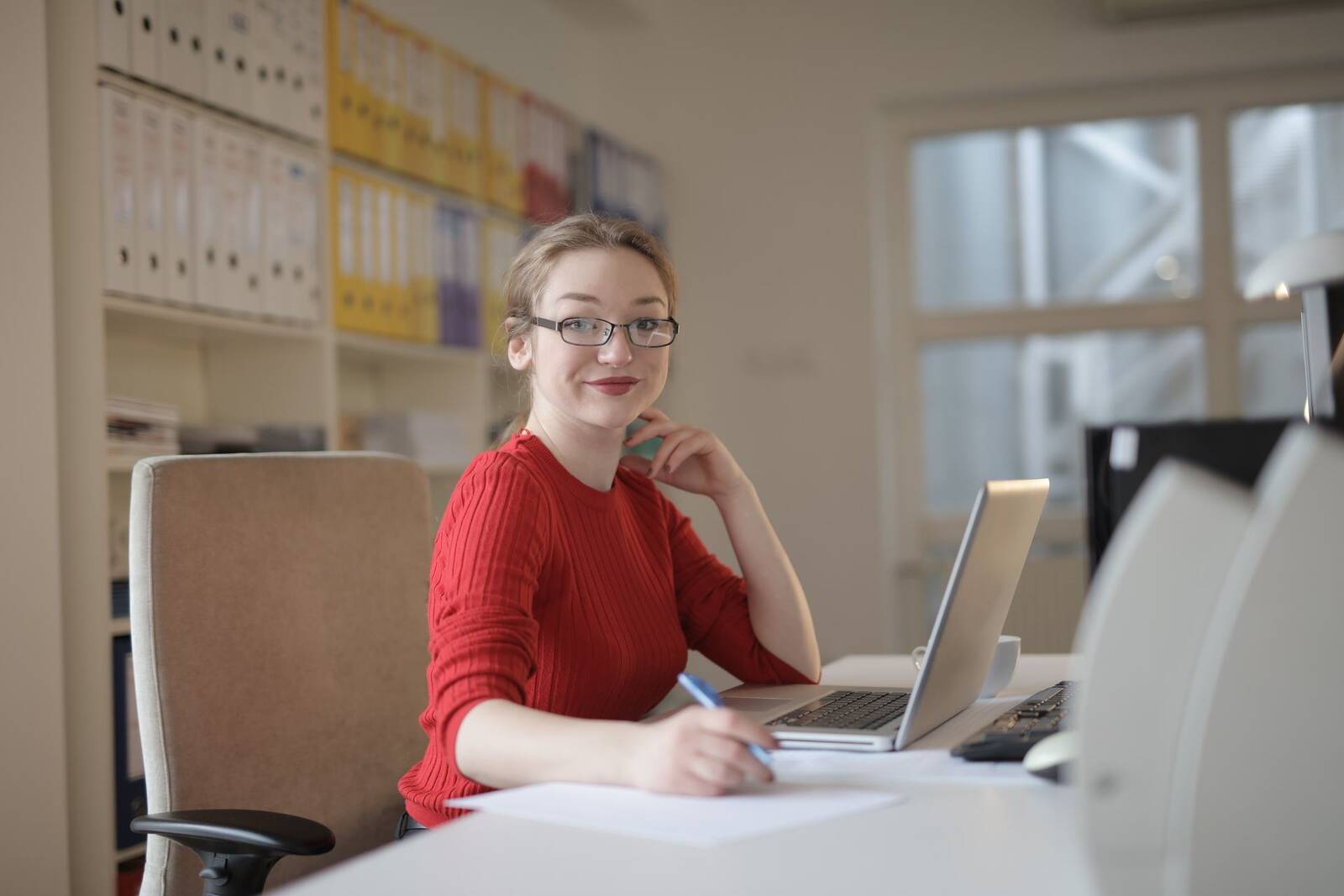 A woman in a red sweater smiles at the camera with a pen in hand, a laptop and papers on the desk in front of her