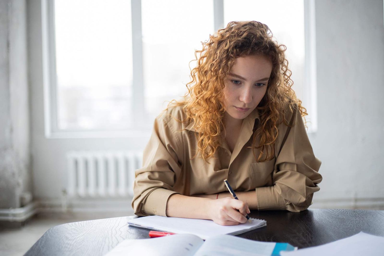 A woman with curly hair sits at a table writing closely in a notebook, an open book and papers spread in front of her