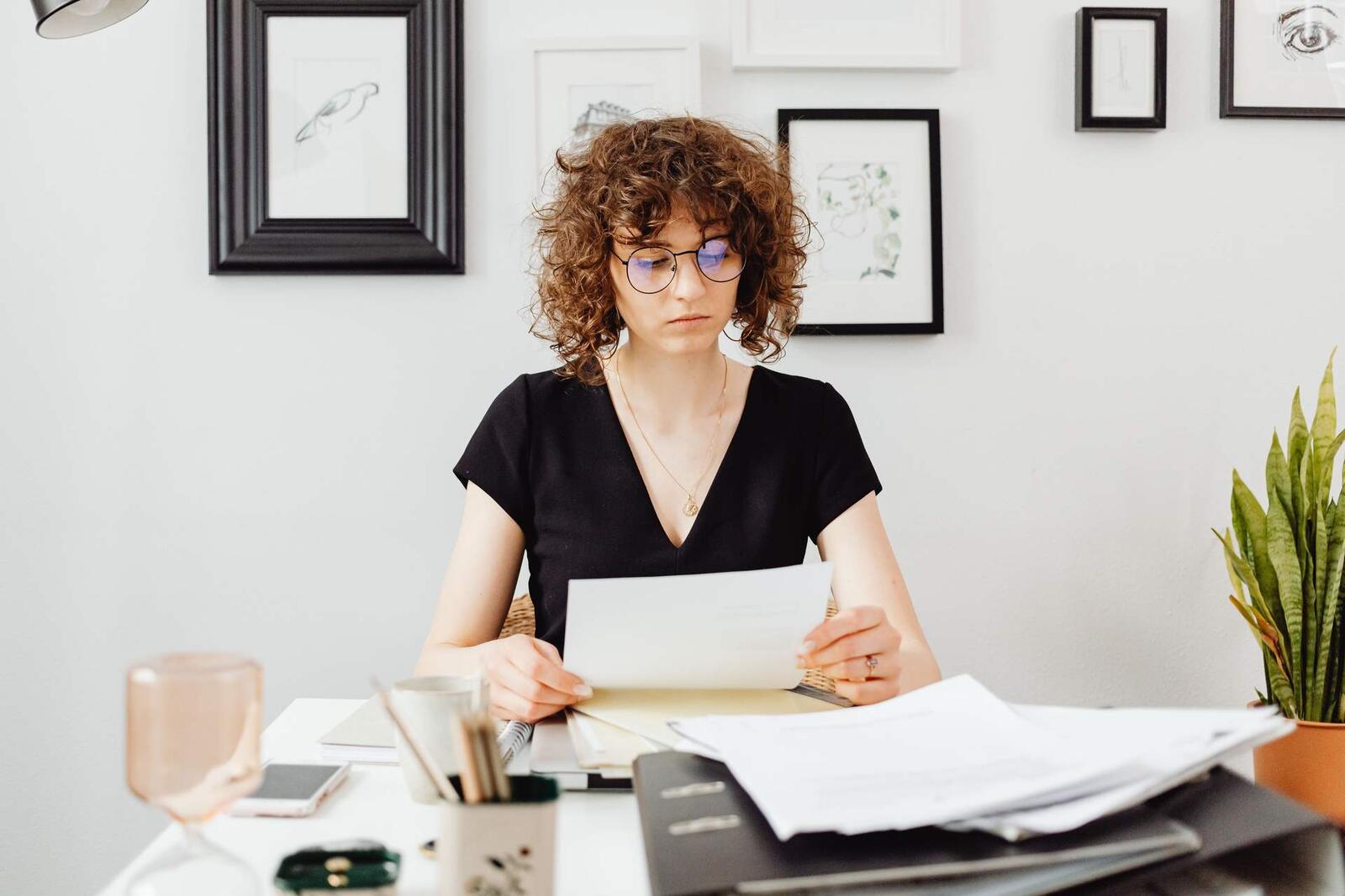 A woman with curly hair and round glasses reads a letter at a white desk with binders and a plant, framed sketches on the wall behind her