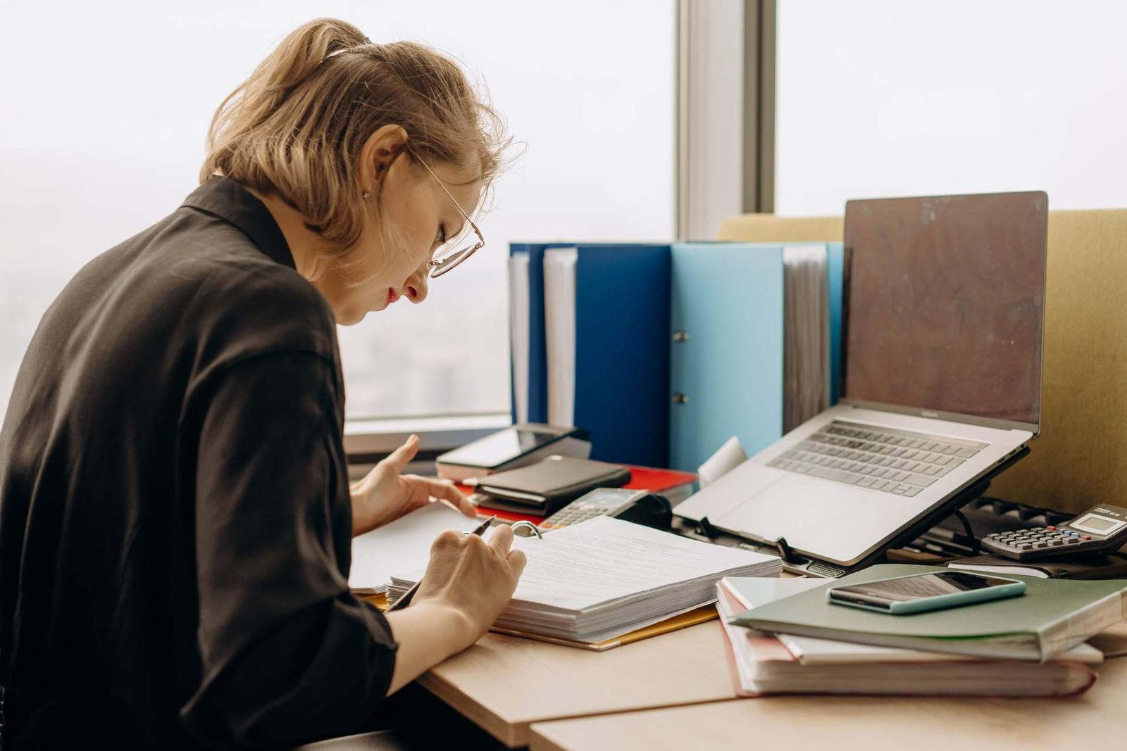 A woman in dark clothing leans over a desk with binders and a laptop, reviewing a thick stack of printed pages