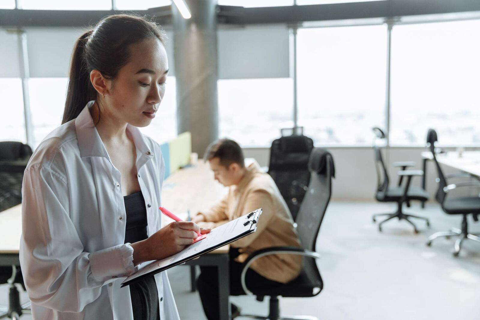 A woman in a white shirt writes on a clipboard in a bright open office, a colleague blurred at a desk in the background
