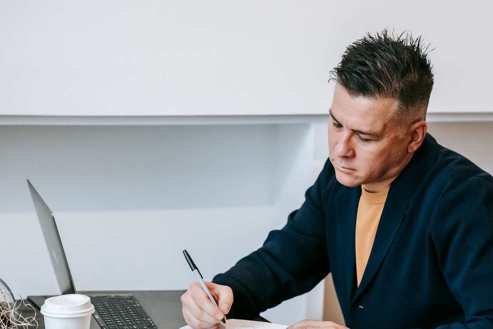 A man writes in a notebook beside an open laptop and a cup of coffee at a low table