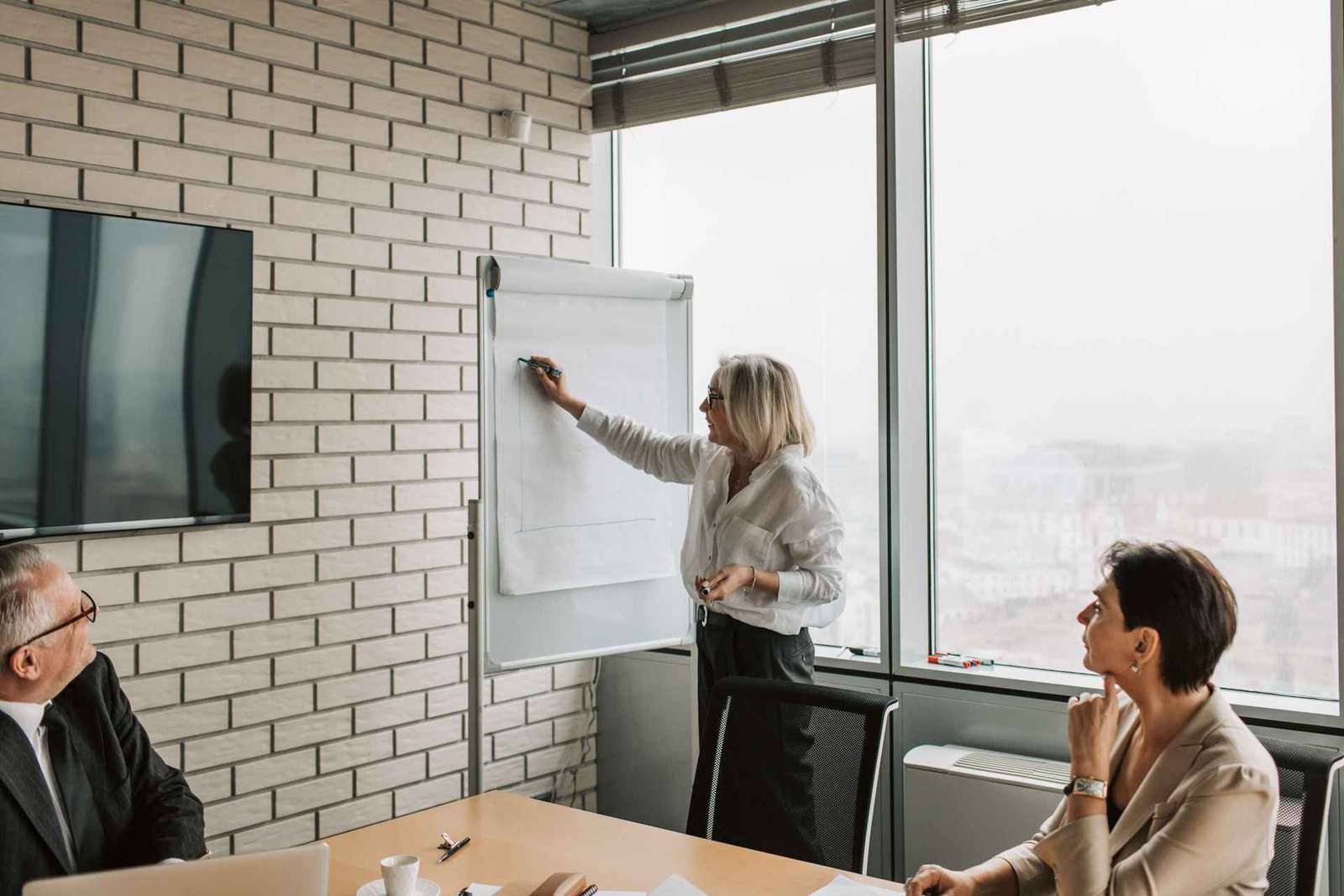 A presenter sketches on a flip chart while two colleagues look on by a window