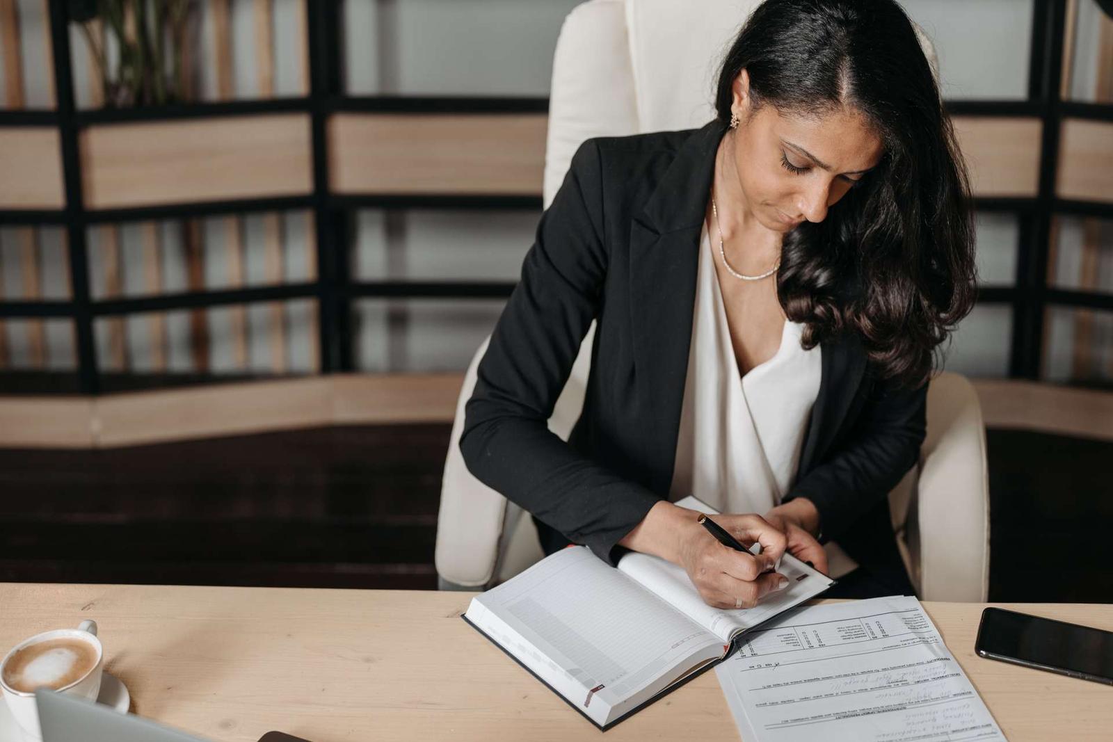 A woman in a black blazer writes closely in a bound notebook at a desk, with a printed form and a coffee cup nearby