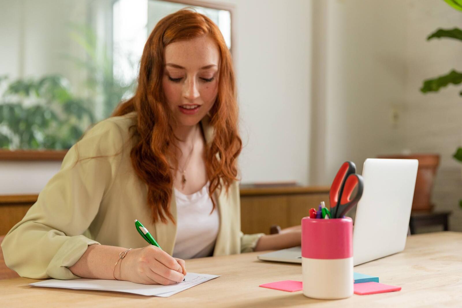 A woman with long red hair, wearing a beige blazer, writes by hand at a desk beside an open laptop and a cup of pens