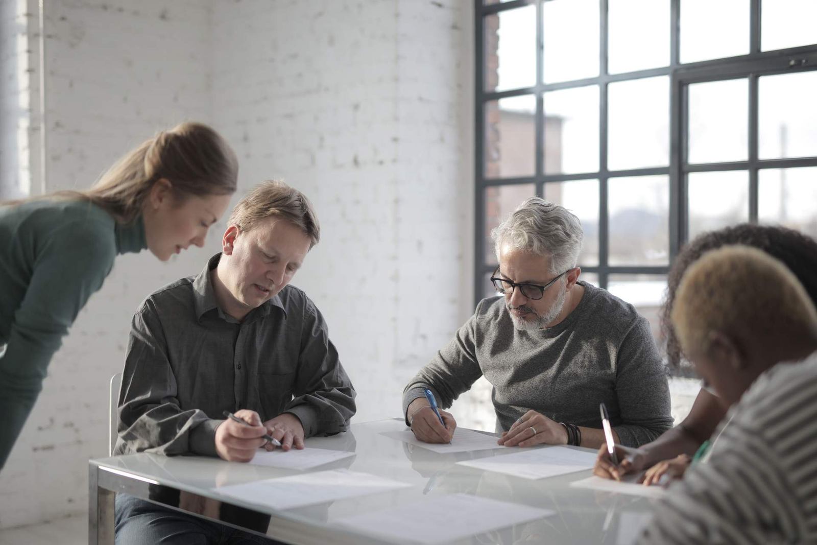 Several adults sit at a table writing on printed documents in a bright, white-brick room with a large window