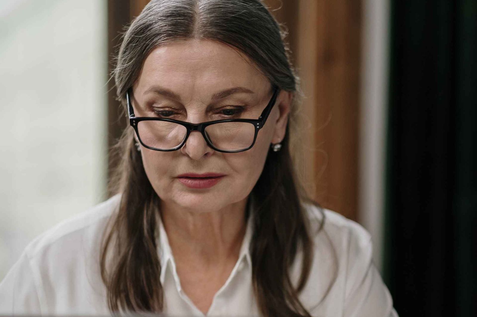 A woman with glasses and gray-streaked hair looks down in concentration, wearing a white blouse, with softly blurred wood paneling and a window behind her