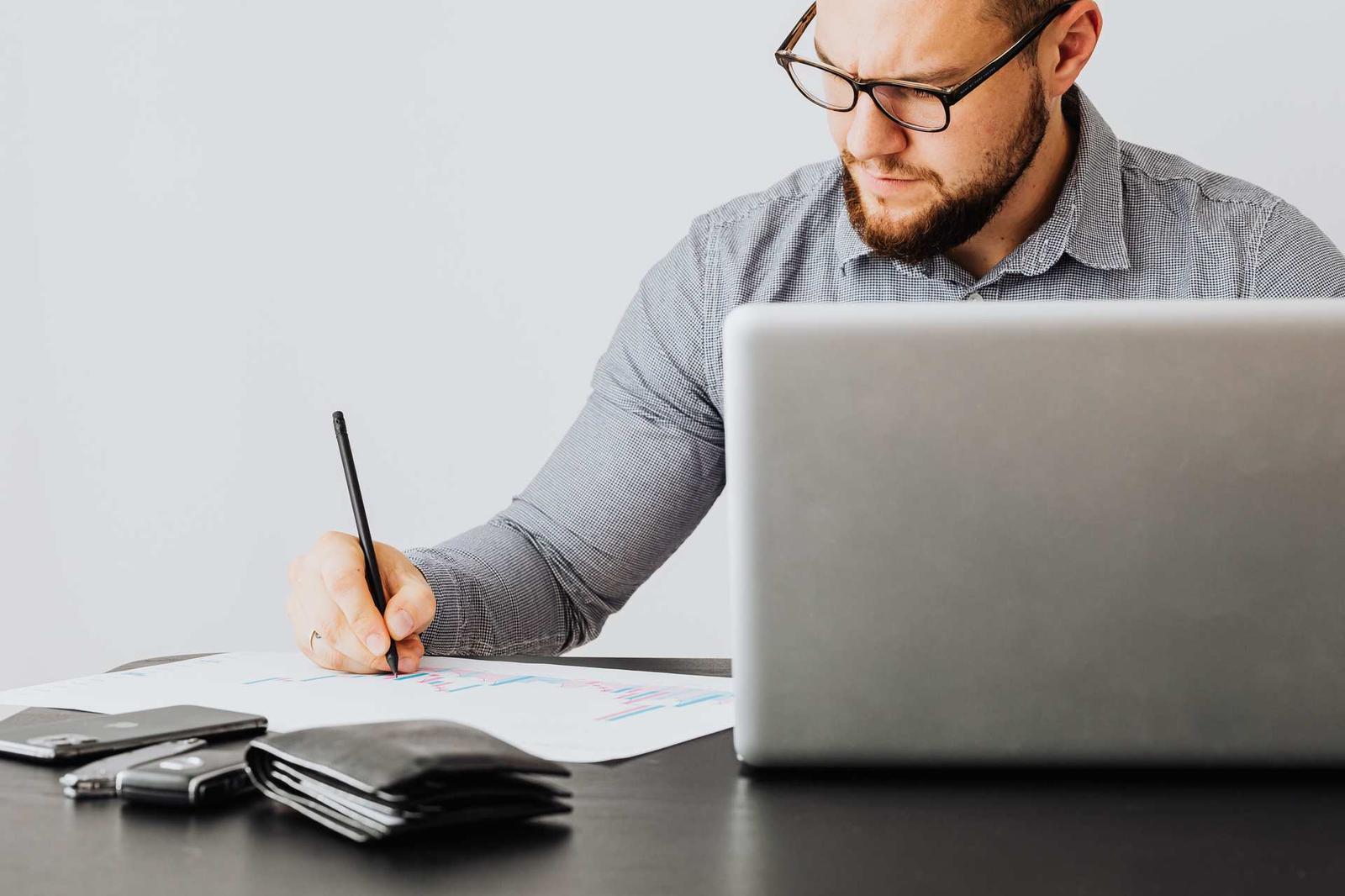 A man wearing glasses annotates a printed chart with a pen while working beside an open laptop