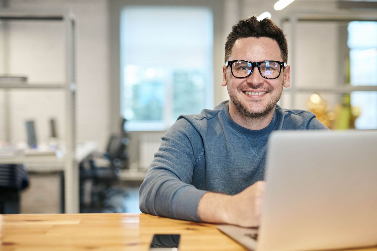 A man in glasses smiles at the camera at a desk with a laptop in a bright office