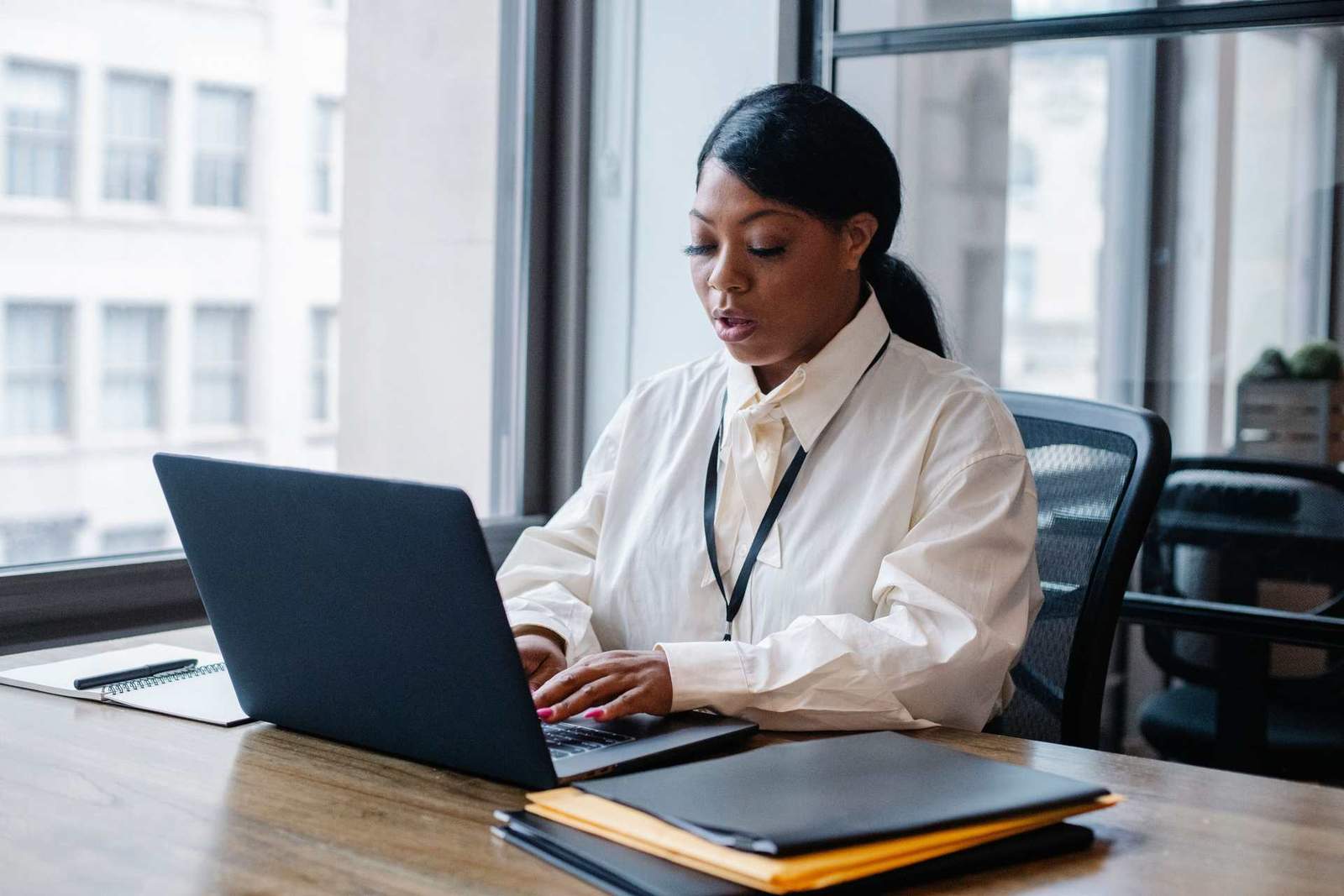 A woman studies her laptop screen closely at a desk in a high-rise office, a badge on a lanyard around her neck