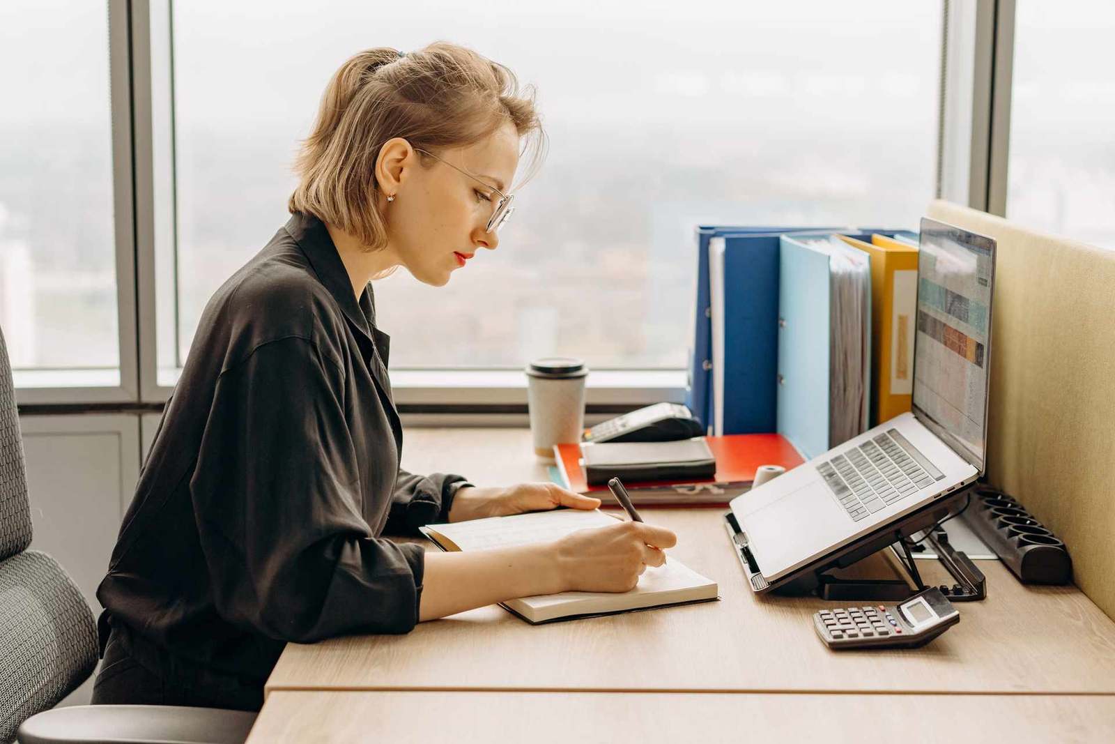 A woman in a black shirt writes in a notebook at a desk with binders and an open laptop, with a city skyline through the window behind her