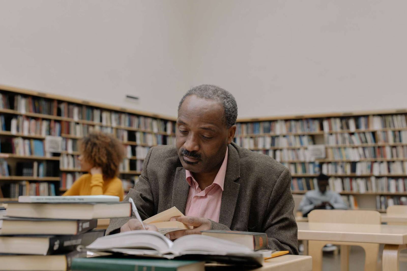 A gray-haired man sits in a library reading closely from a book, a stack of reference volumes beside him