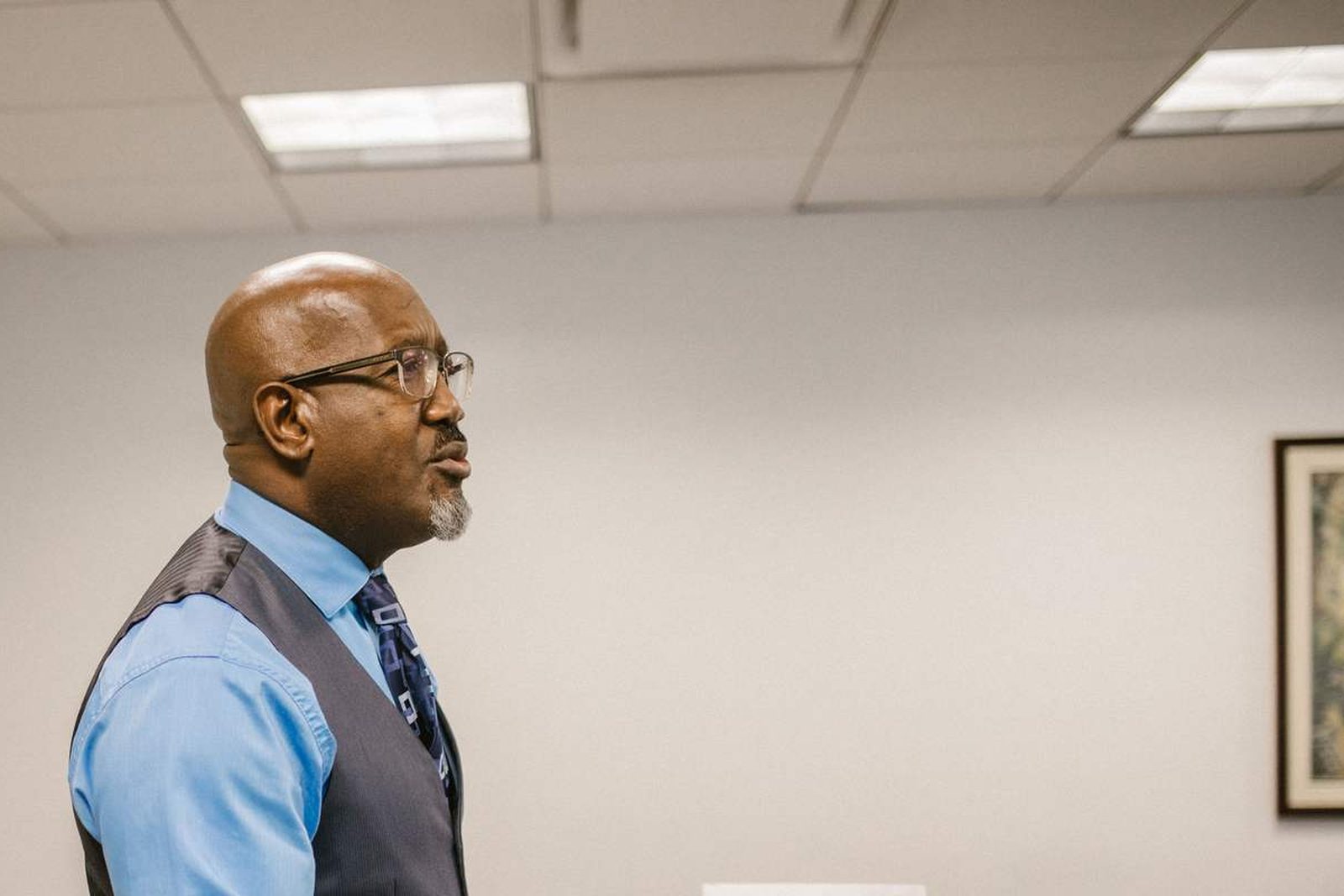 A man in a vest and tie stands in profile against a plain office wall, glasses on, mid-sentence