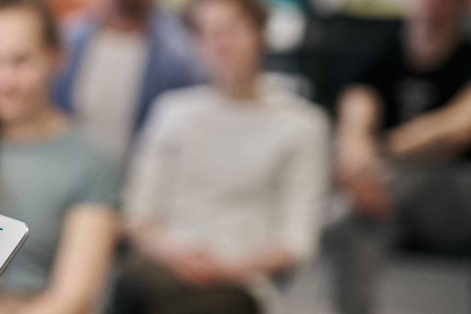 A blurred seminar audience seen past an unfocused foreground, a hand and the edge of a tablet visible at the left