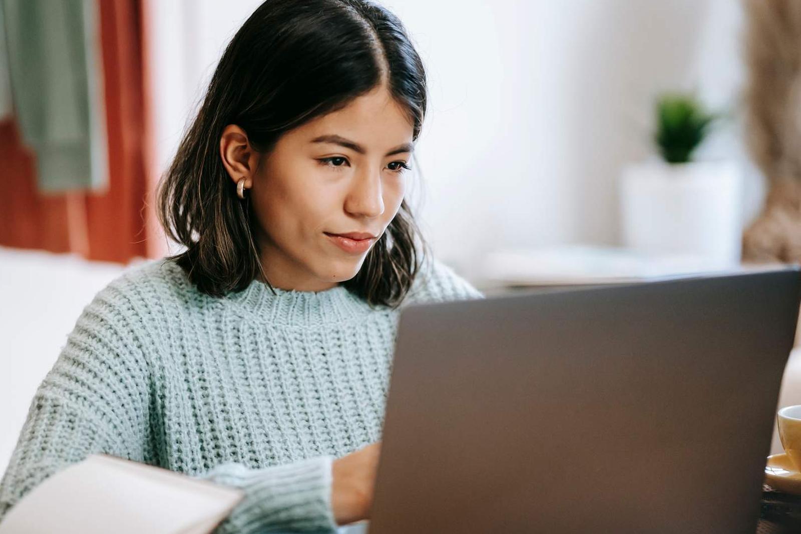A woman in a light green sweater looks intently at an open laptop while working at a desk
