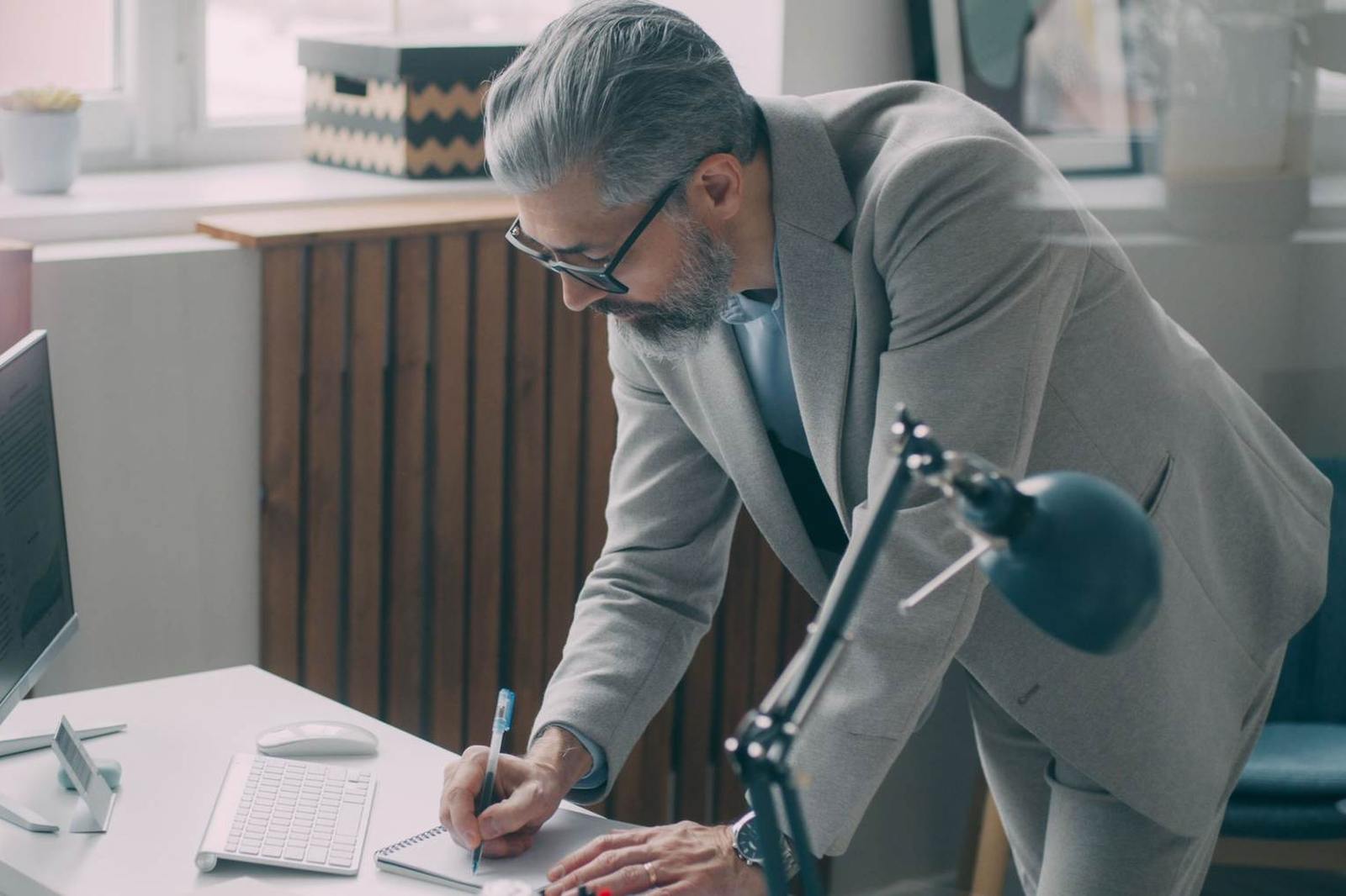 A gray-haired man in a blazer leans over a standing desk, writing in a notebook beside a monitor and an adjustable desk lamp