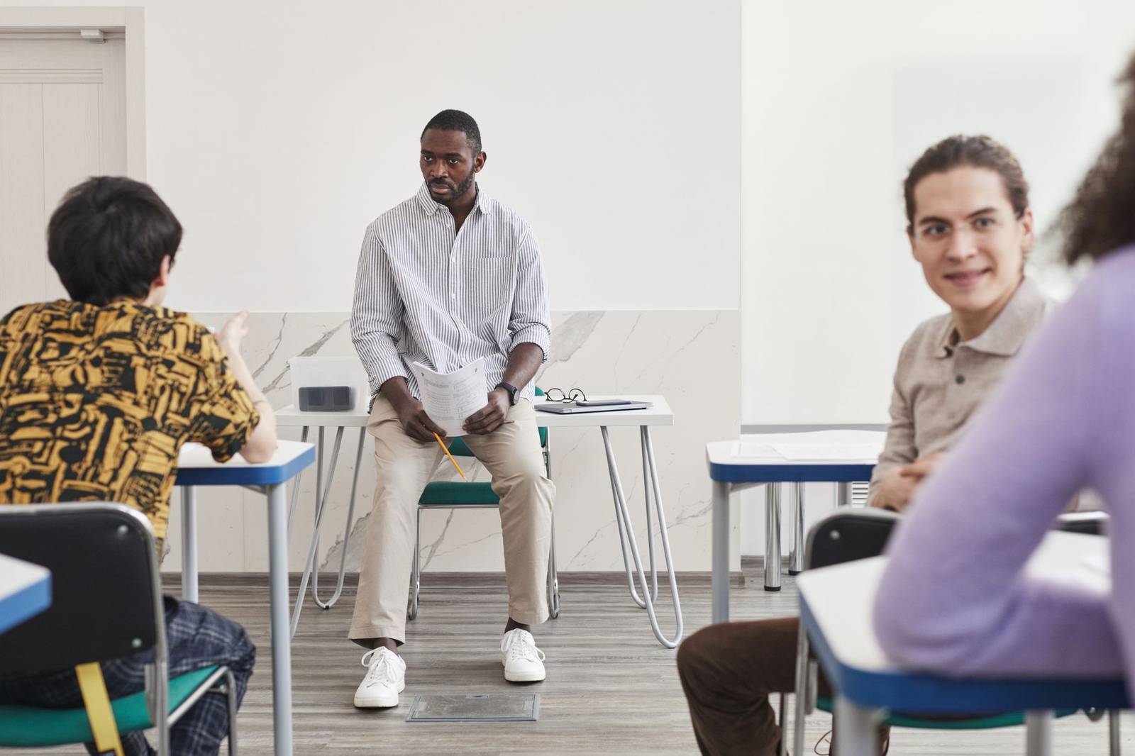 A man stands at the center of a semicircle of classroom desks, holding a printed handout, while three seated participants turn to listen during a group discussion