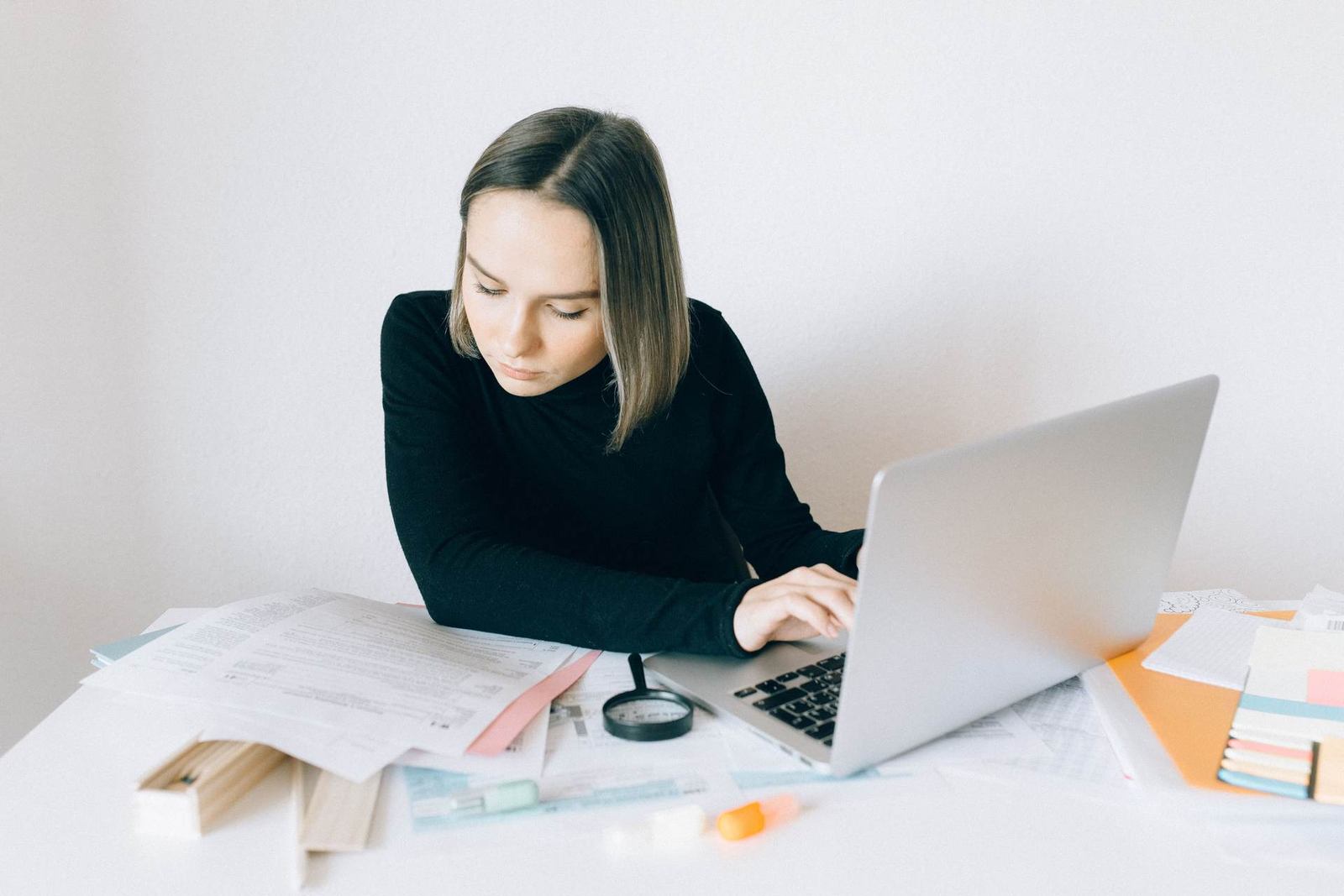 A woman in a black top leans over a laptop at a desk covered in papers, typing with one hand