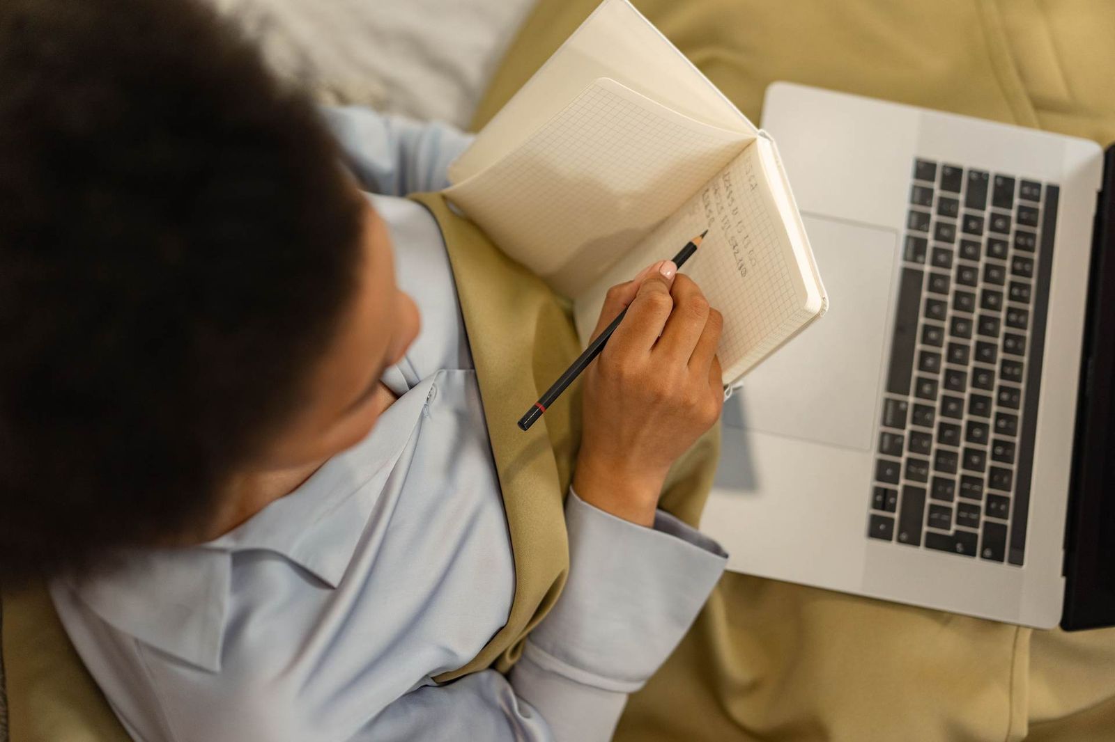 An overhead view of a person writing notes in a spiral notebook beside an open laptop