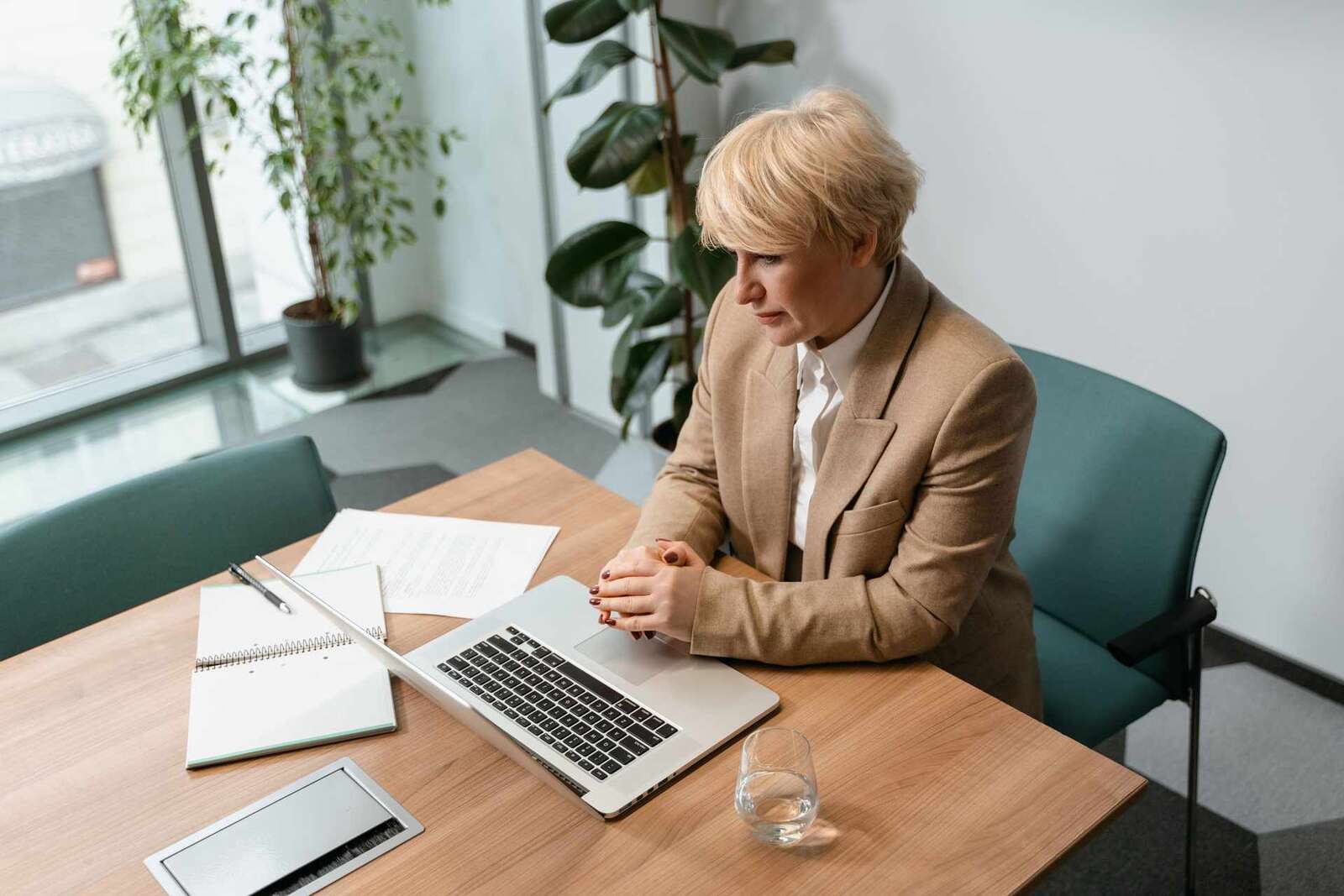 A woman in a tan blazer sits with hands clasped, looking intently at a laptop in a meeting room