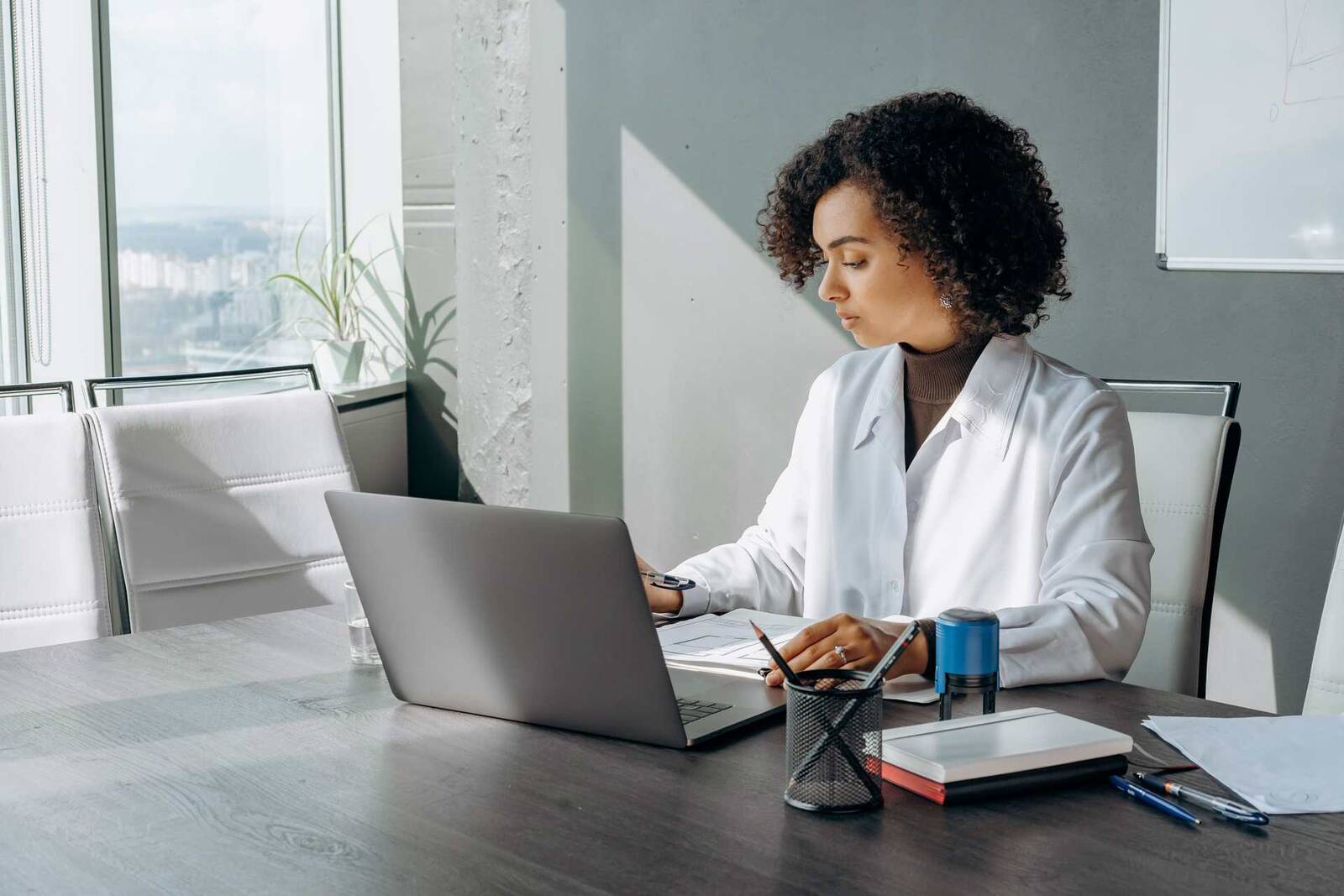 A woman in a white blazer reviews printed pages beside an open laptop in a bright conference room