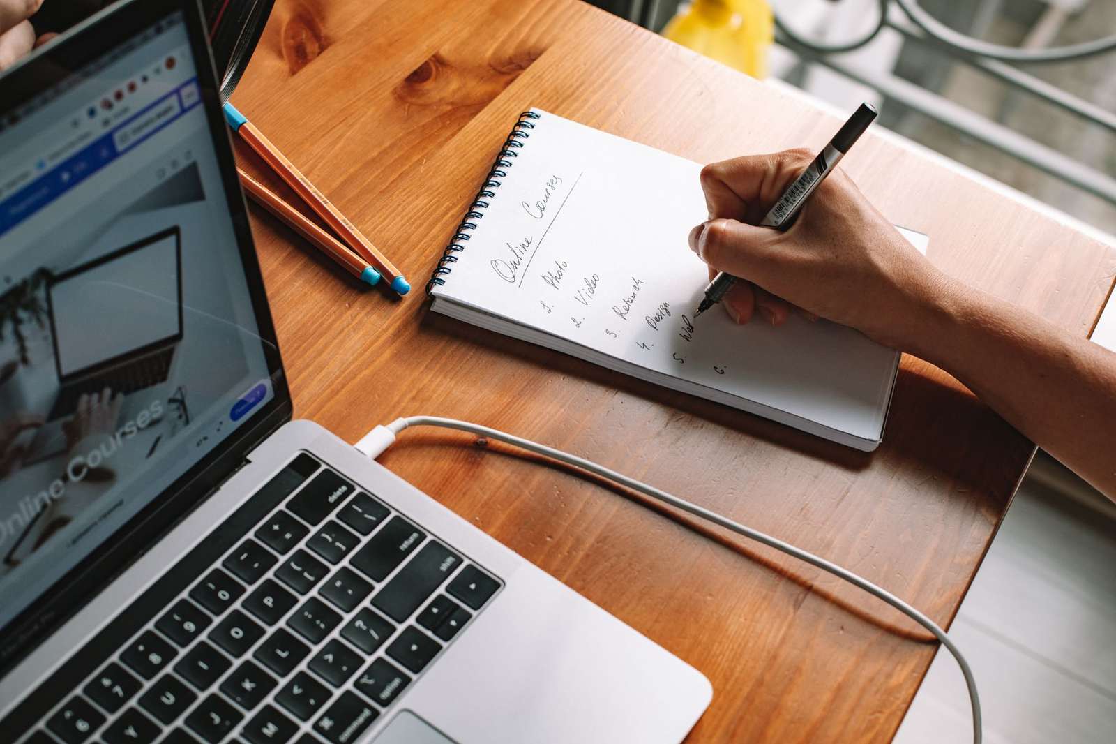 A hand writes a numbered list in a spiral notebook beside an open laptop, with colored pencils on a wooden desk
