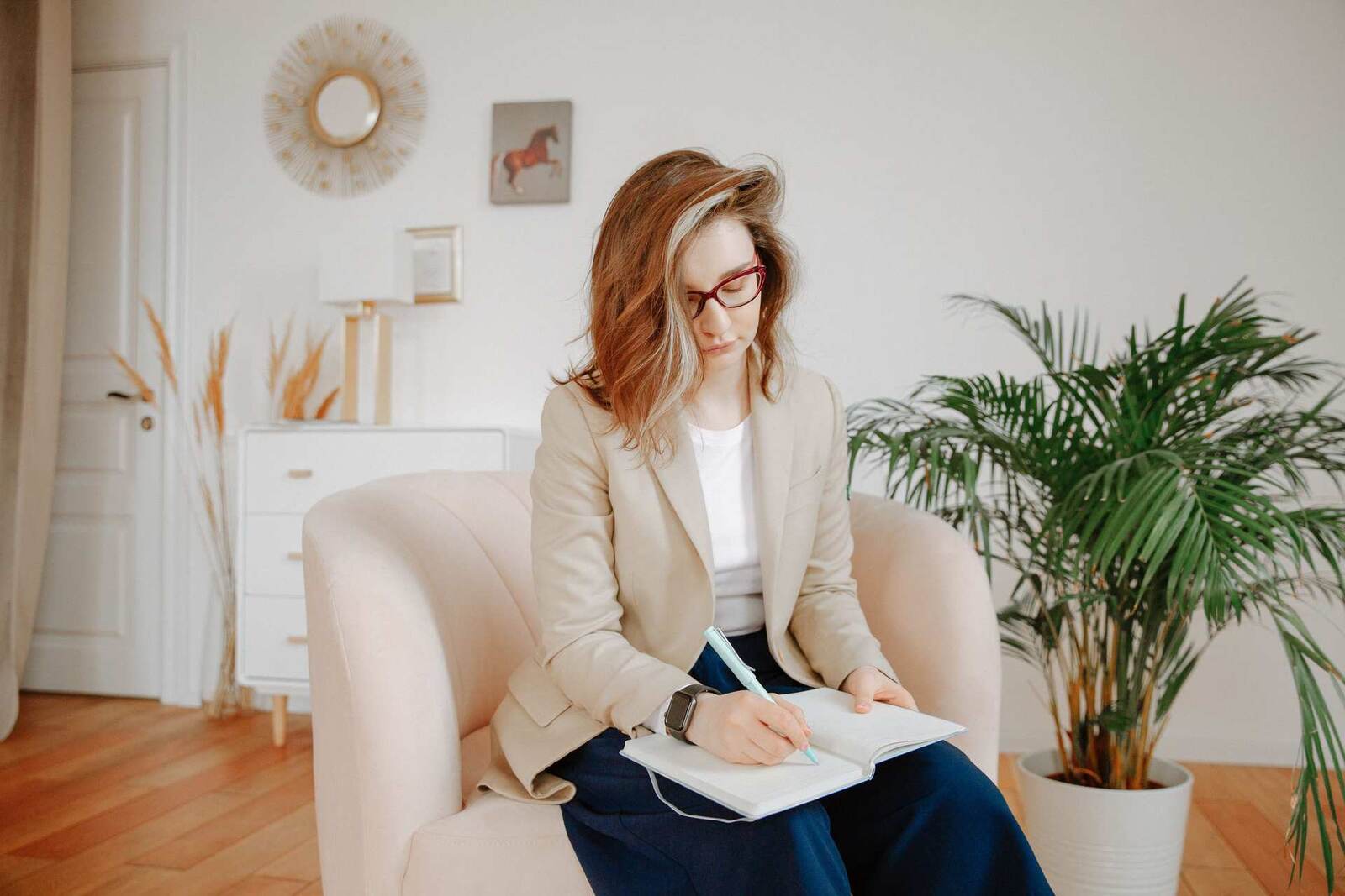 A woman in a beige blazer and red glasses writes in a notebook while seated in an armchair in a bright living room, a palm plant beside her
