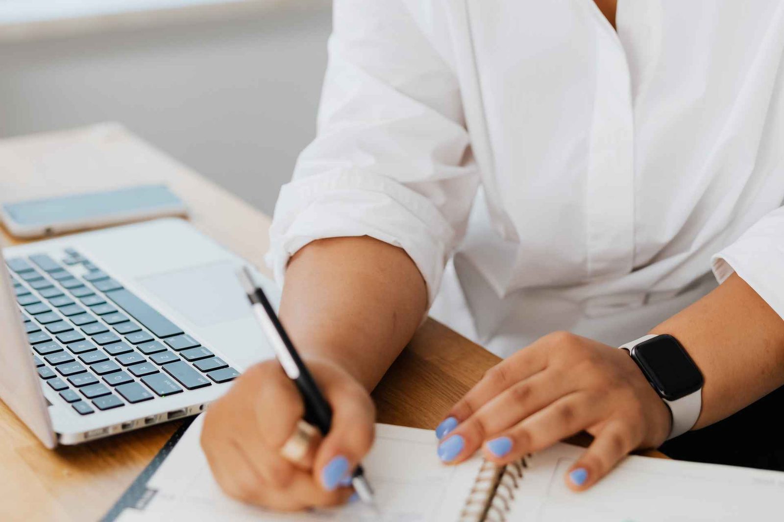 A close view of hands writing in a spiral notebook at a desk beside an open laptop and a phone, no face visible