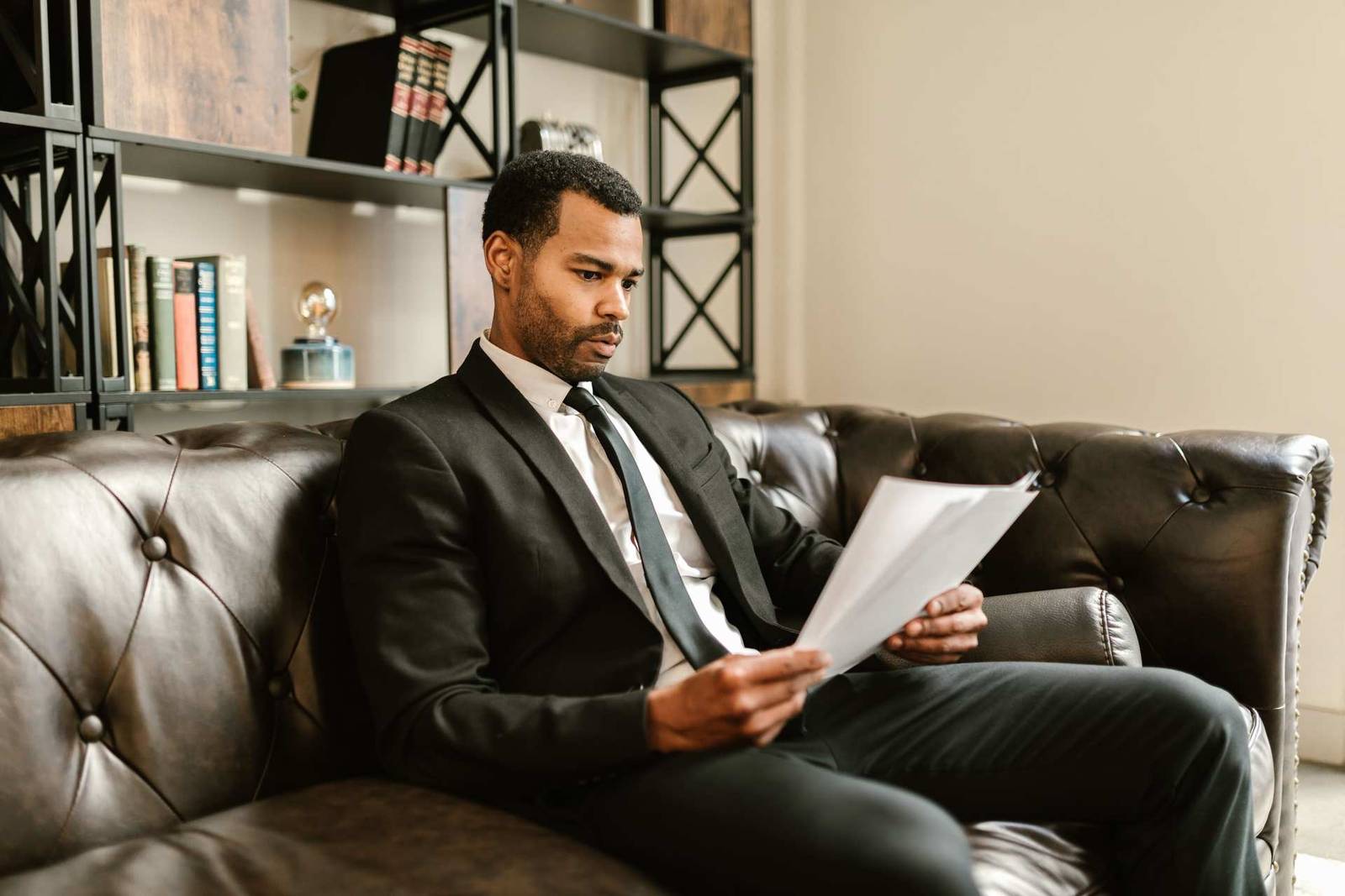 A man in a black suit sits on a leather couch reading a printed document, with a bookshelf of books in the background