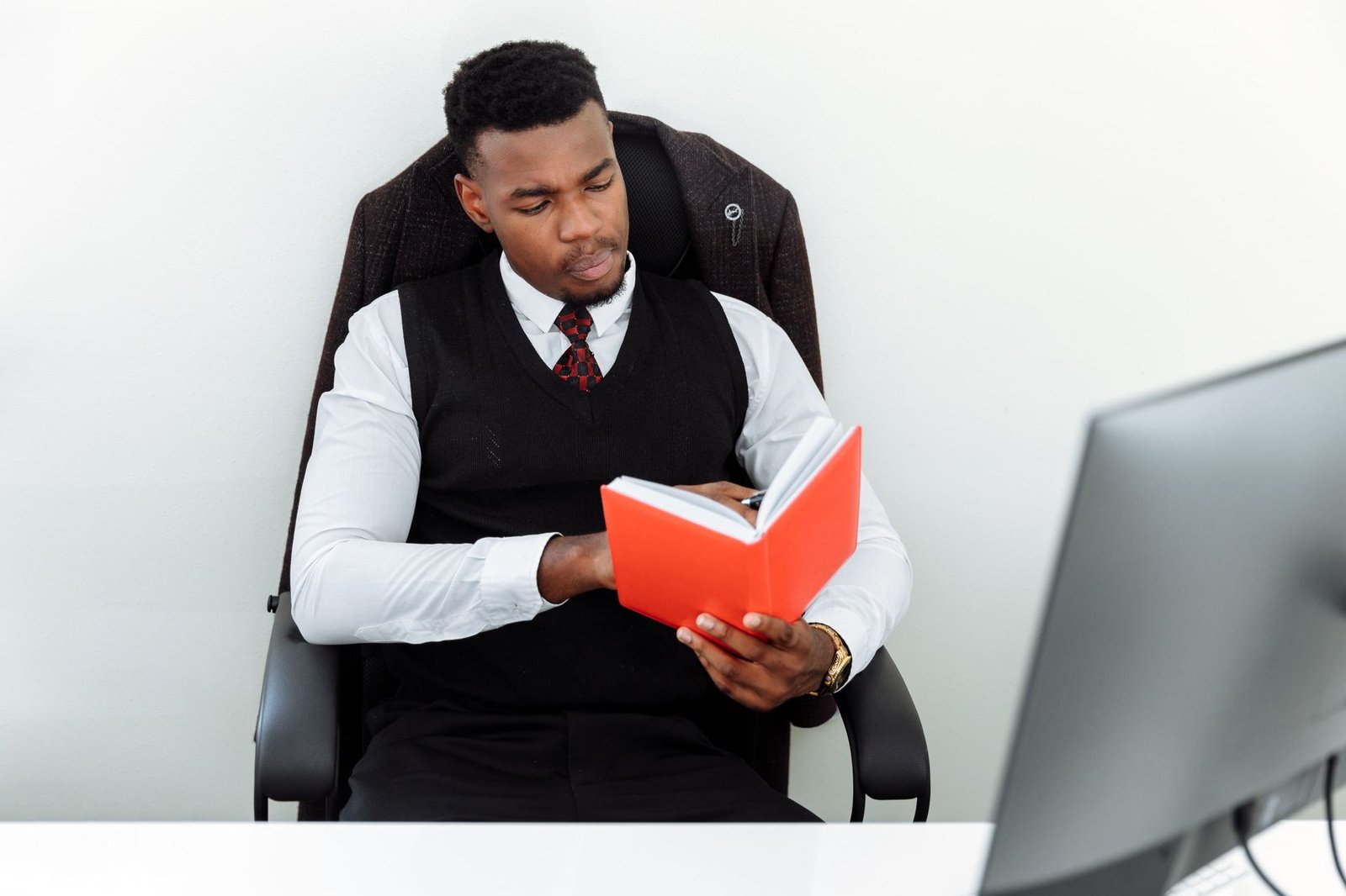 A man in a vest and tie sits reading closely from an open notebook at his desk beside a computer monitor