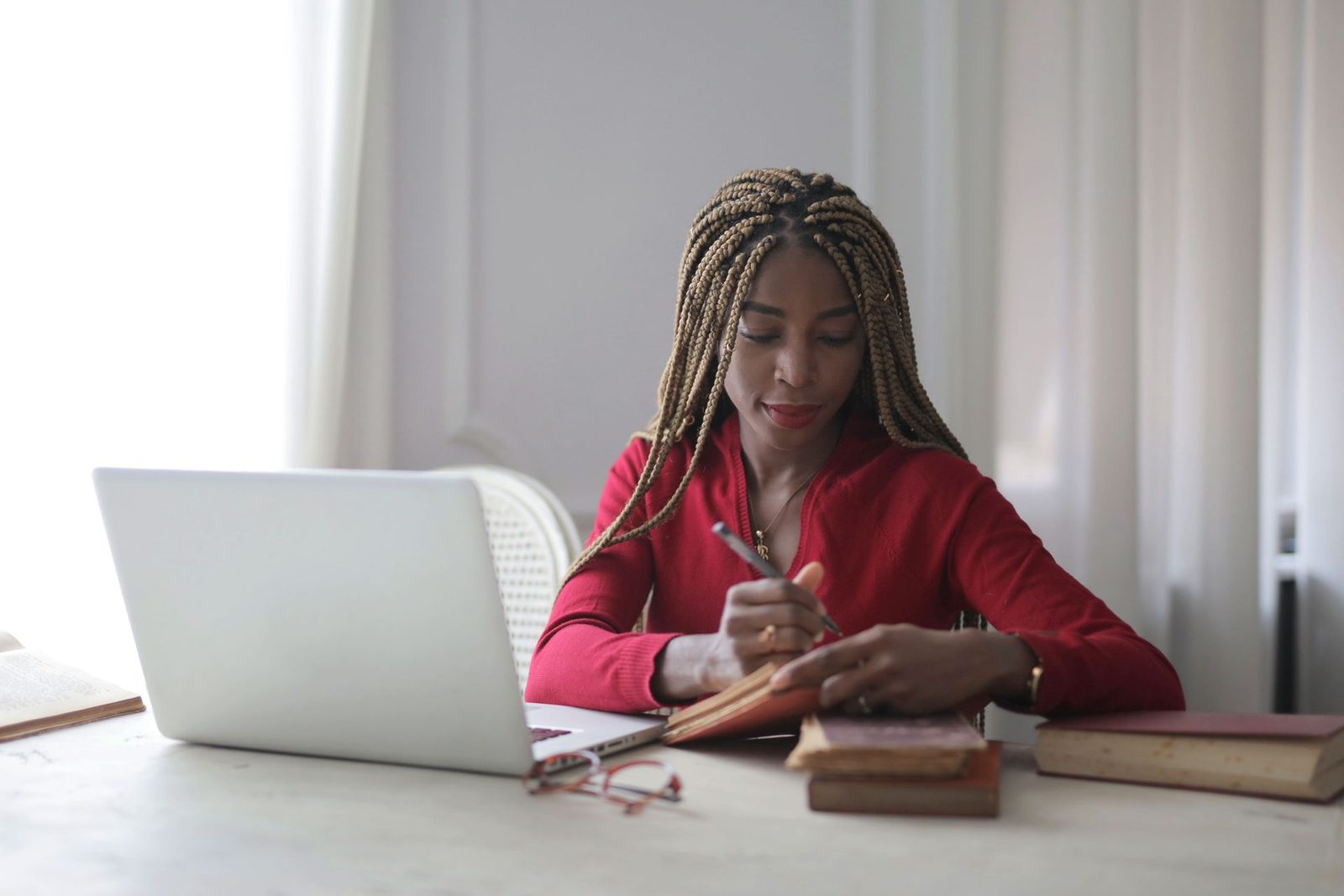 A woman in a red sweater writes in a notebook at a desk, an open laptop and a stack of old books beside her