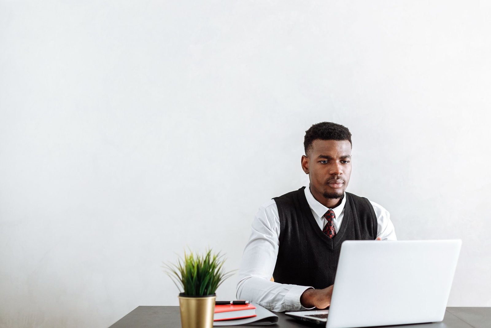 A man works closely at a laptop against a plain white office wall, a small potted plant on the desk beside him