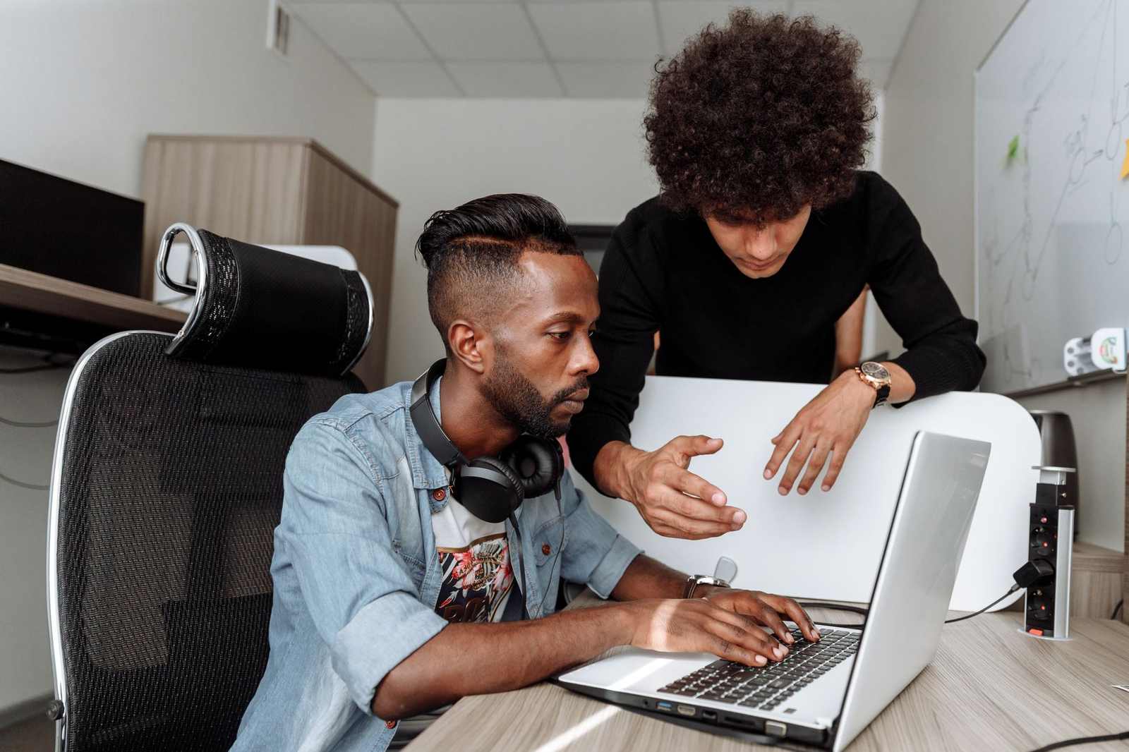 Two colleagues look closely at a laptop screen together at a desk, one seated and typing while the other leans in and points