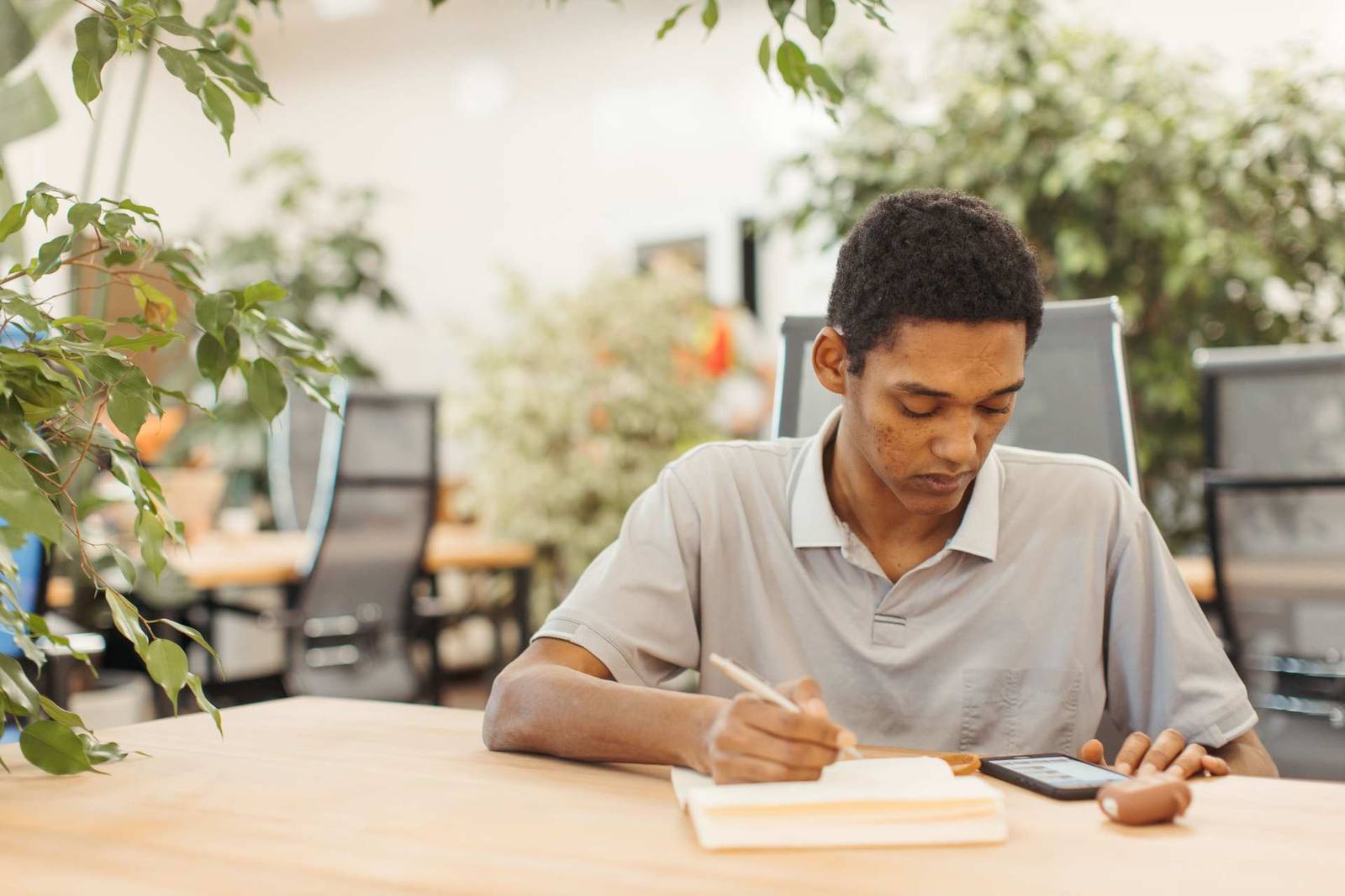A man writes in a notebook at a wooden table surrounded by office plants