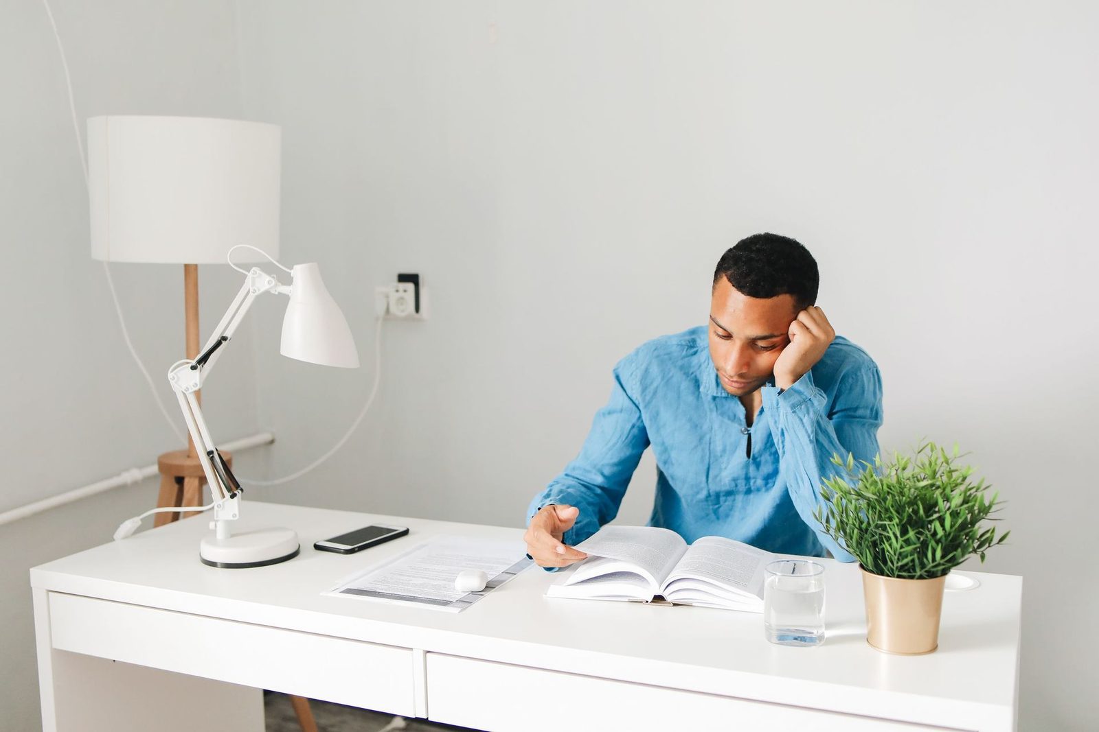 A man in a blue shirt reads an open book at a white desk beside a lamp and a small potted plant