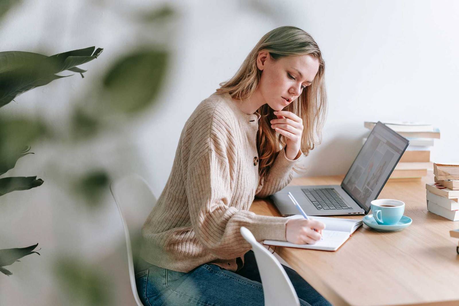 A woman sits at a desk with an open laptop, writing in a notebook beside a cup of coffee and a stack of books