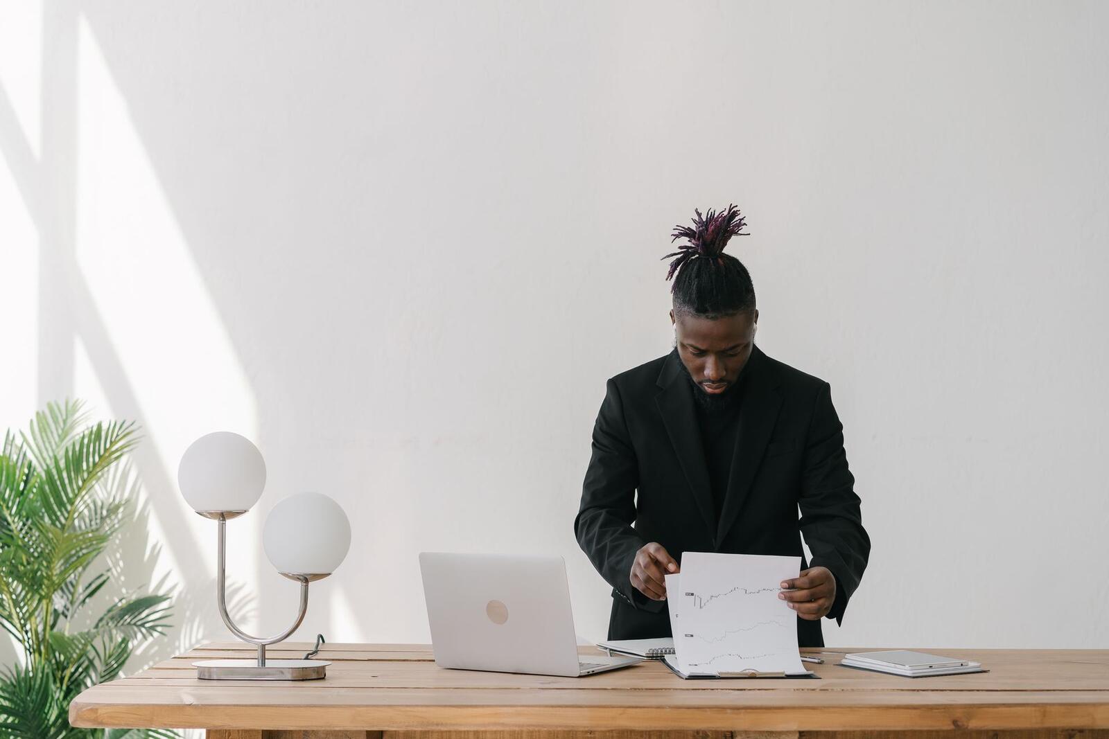 A man in a black blazer reviews printed line charts at a desk beside an open laptop