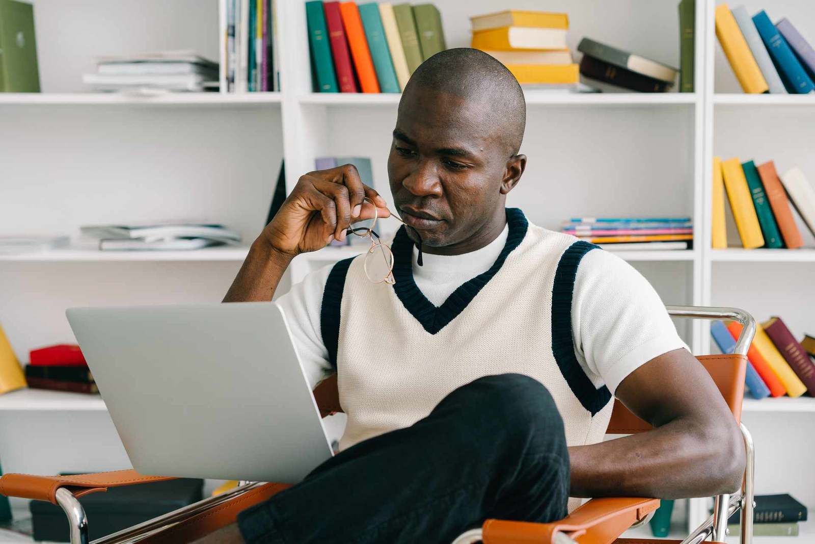 A man takes off his glasses while studying a laptop screen in a bright, book-lined room