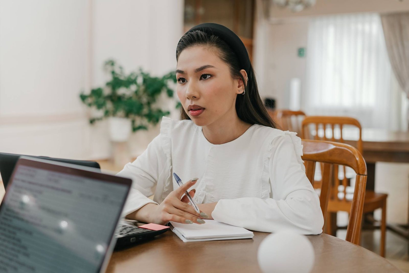 A woman in a white blouse holds a pen at a desk, an open laptop and notebook in front of her